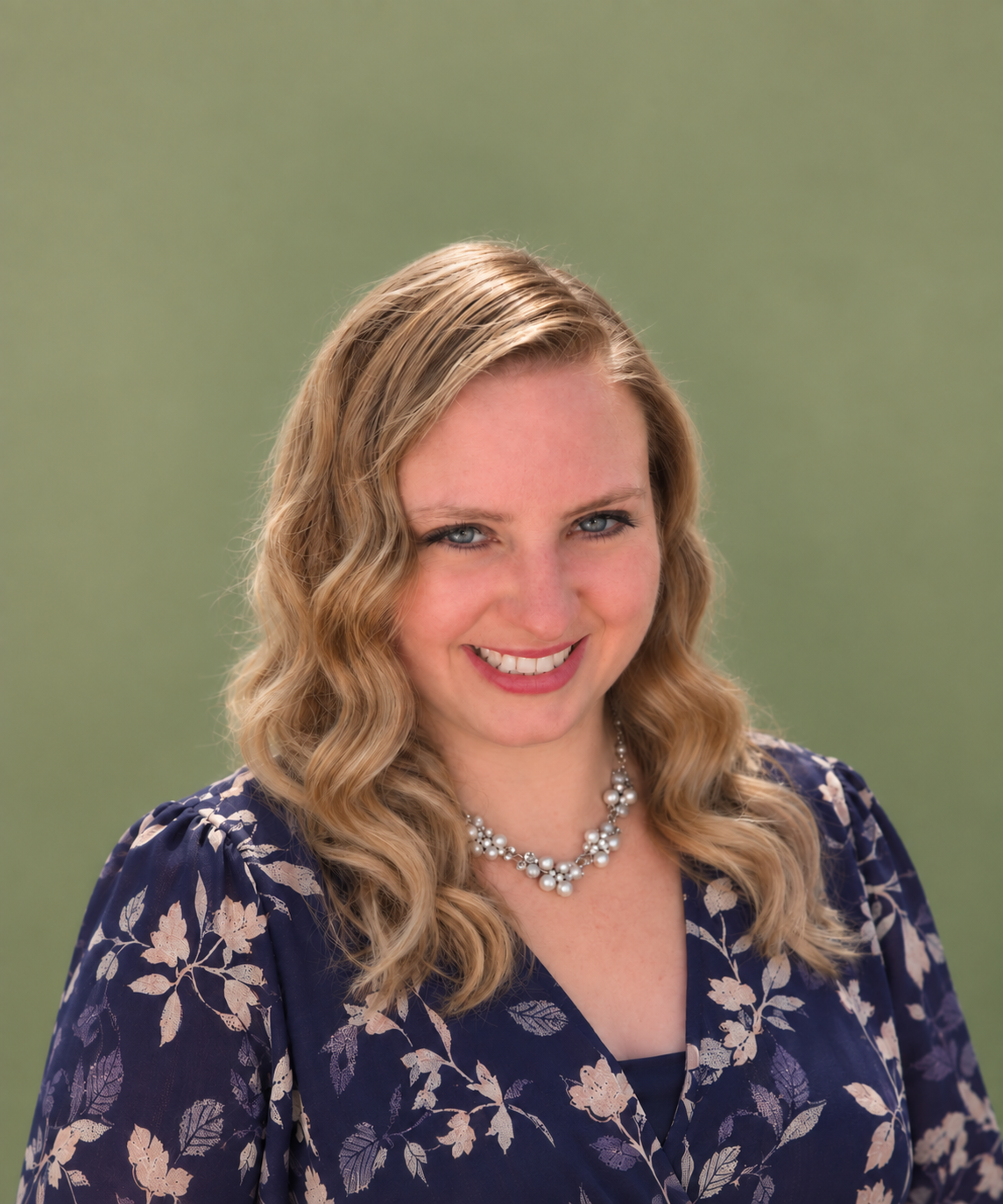 A woman with long, wavy blonde hair, blue eyes, and a bright smile, wearing a navy blue floral dress and a pearl necklace, posing outdoors with a blurred green background.