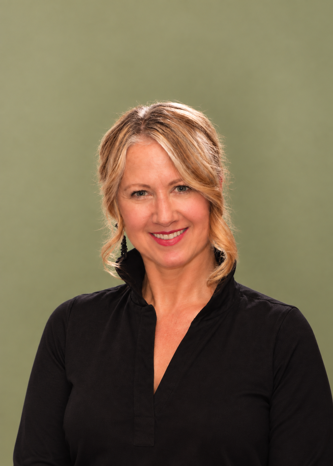A smiling woman with blonde hair styled in loose curls, wearing a black top with a high collar, standing against a solid green background.