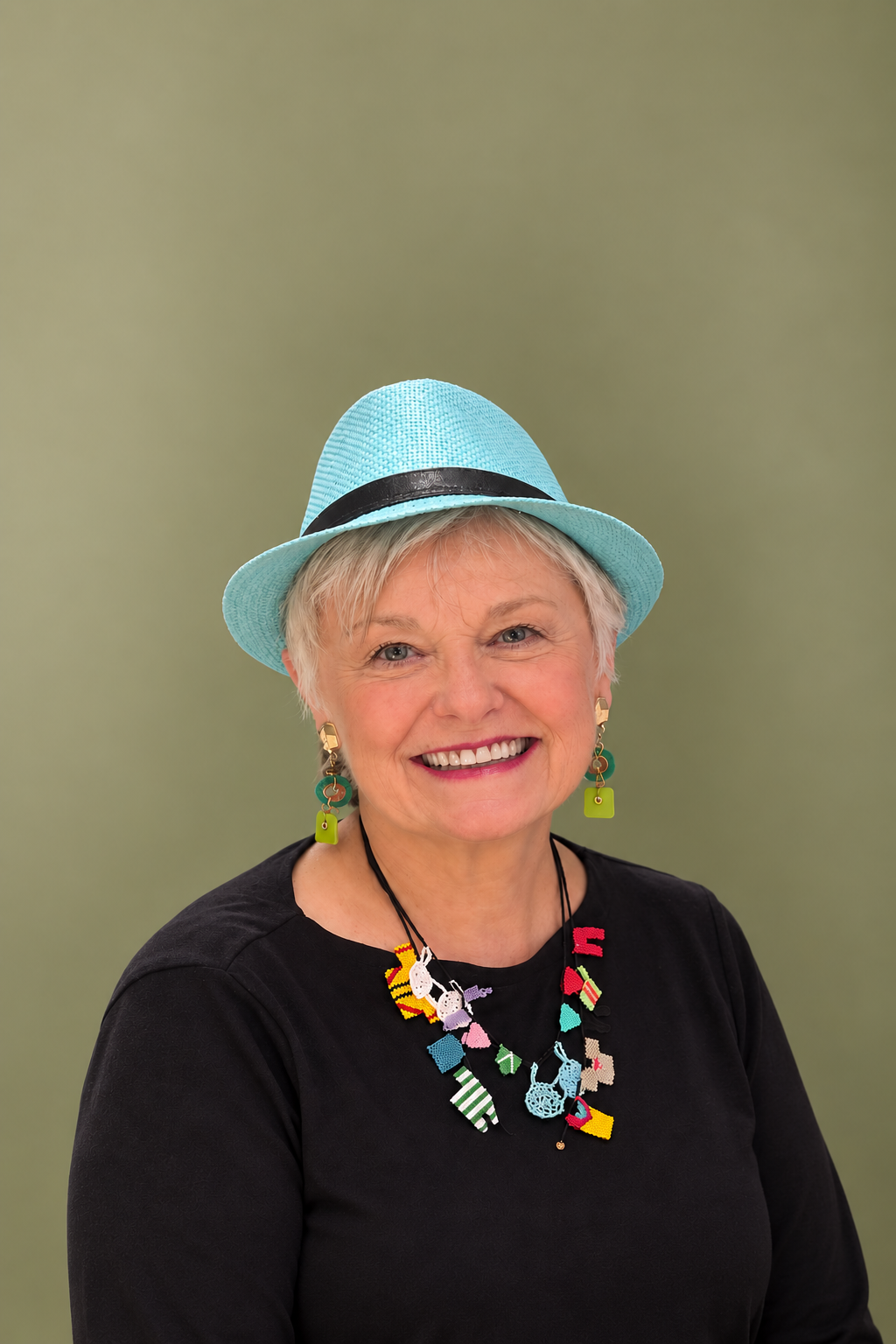 A smiling woman wearing a light blue hat with a black band, colorful earrings, and a necklace with yarn and fabric candy shapes, against a green background.