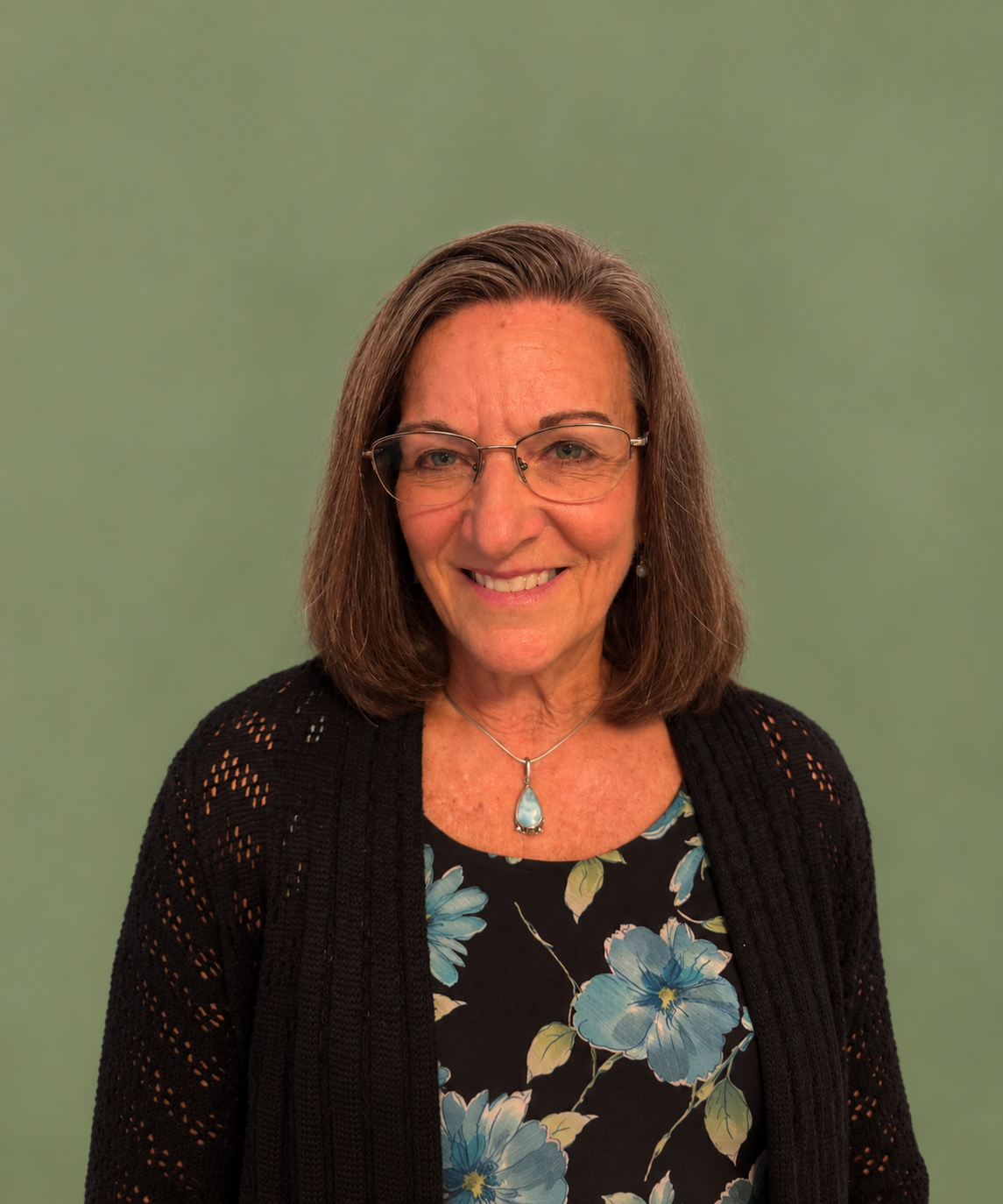 A smiling woman with shoulder-length brown hair, wearing glasses, a black floral top, a black cardigan, and a turquoise pendant necklace, standing against a plain green background.