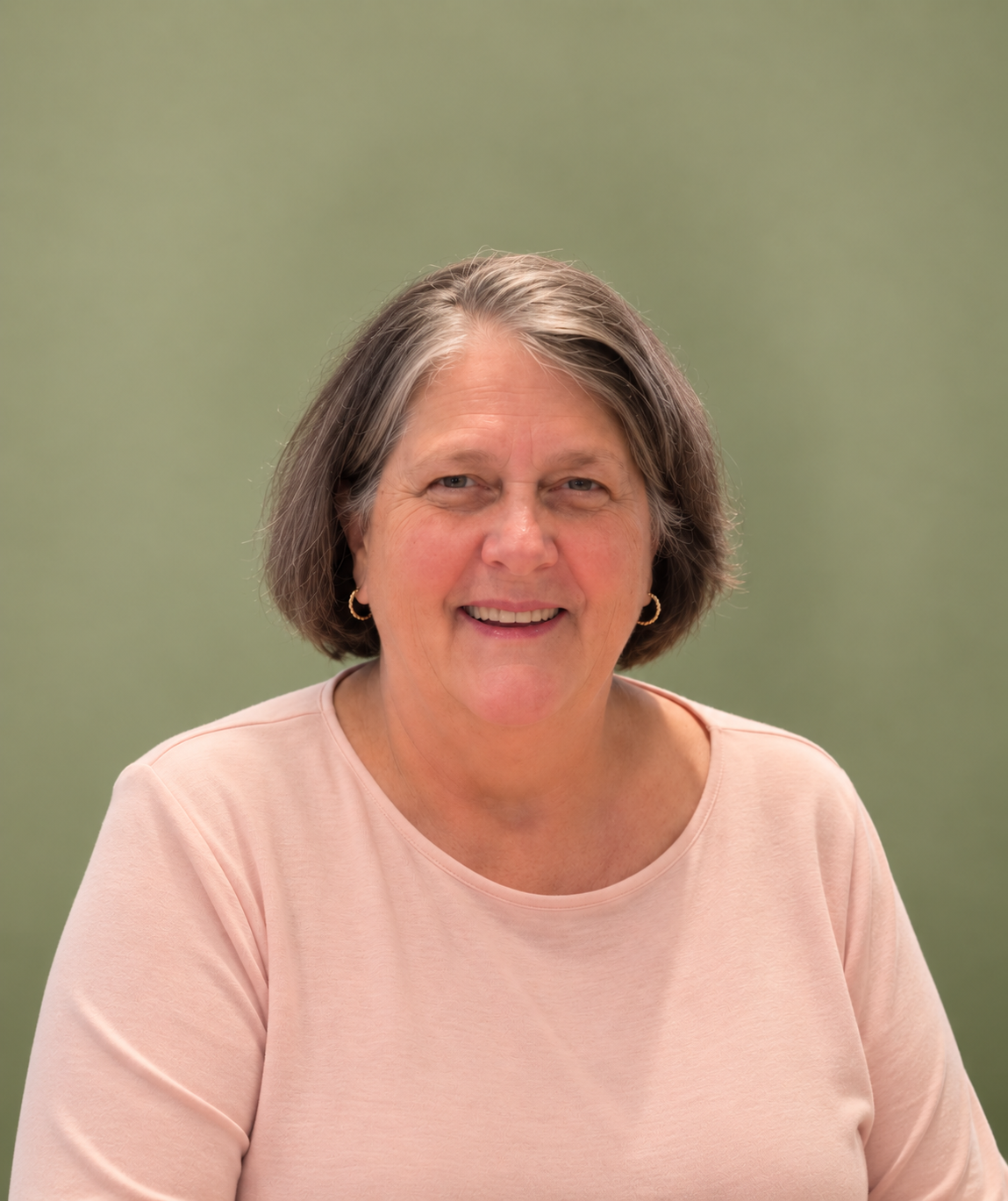 A smiling woman with shoulder-length gray hair, wearing a light pink top and small hoop earrings, against a blurred green background.
