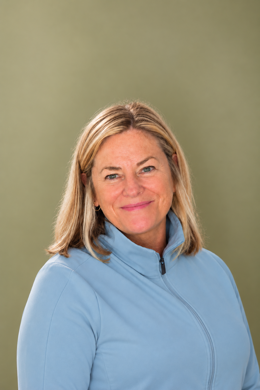 A woman with blonde hair wearing a light blue zip-up jacket smiling against a plain green background.