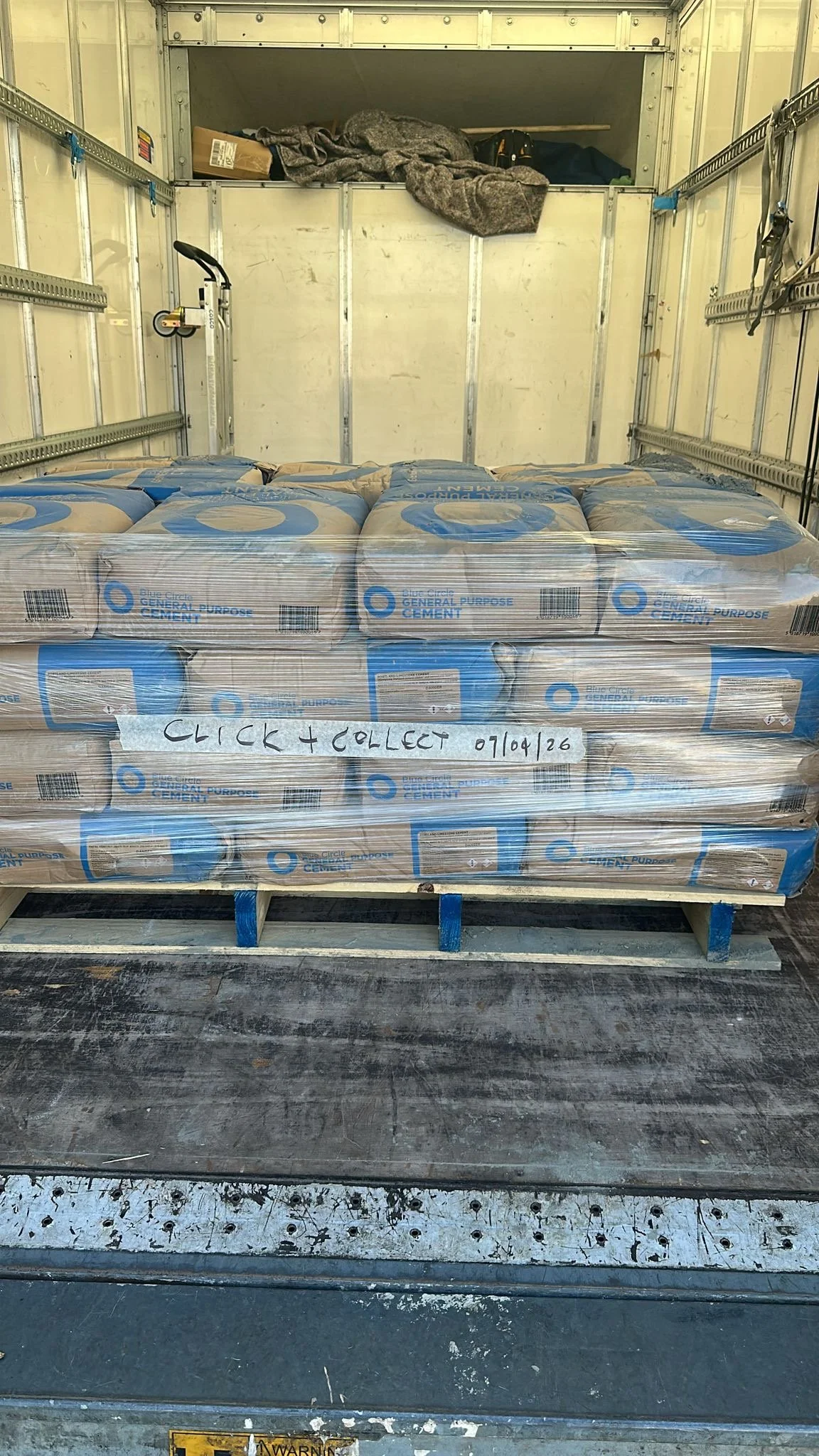 A pallet of bags of general purpose cement wrapped in plastic, with a handwritten sign reading 'Click & Collect 01/09/26' attached to the pallet, inside a delivery truck.