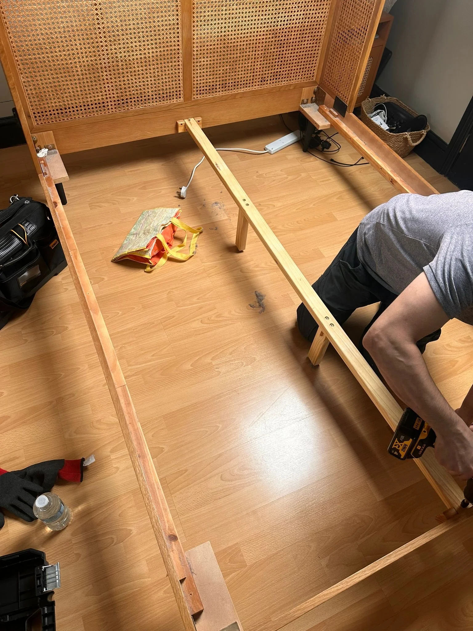 Person assembling a wooden bed frame on a hardwood floor. Tools, gloves, and a water bottle are nearby.
