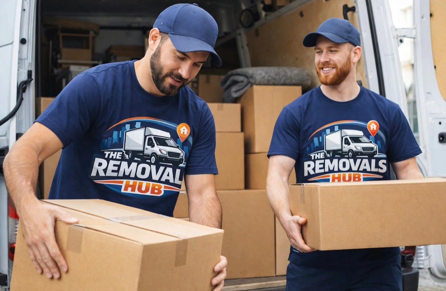 Two men in navy blue T-shirts and caps with 'The Removal Hub' logo pack boxes into a white van, working together during a moving service.
