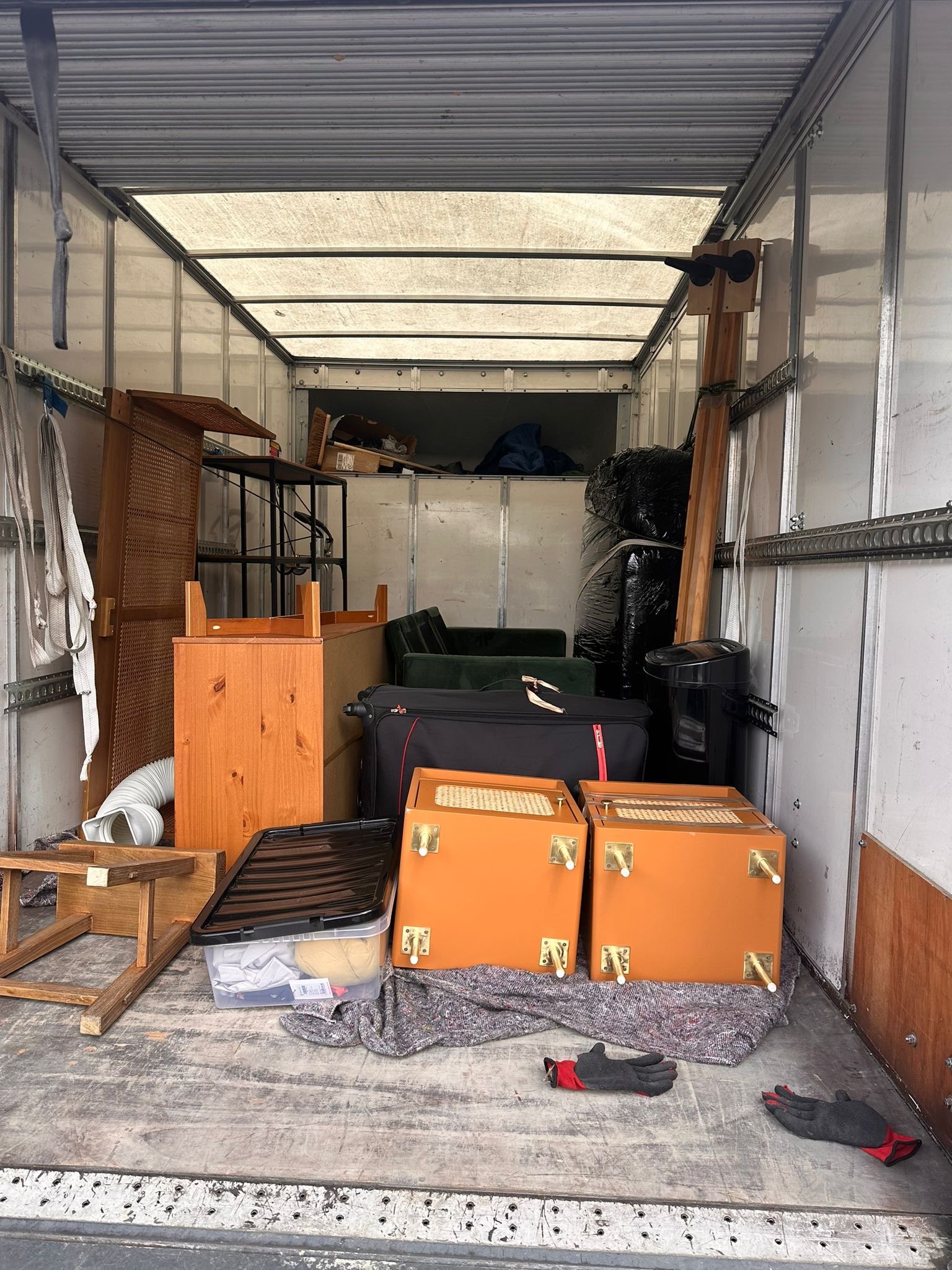 Interior of a moving truck filled with various furniture pieces including a small table, a black suitcase, wooden cabinets, and wrapped items, with work gloves on the floor.