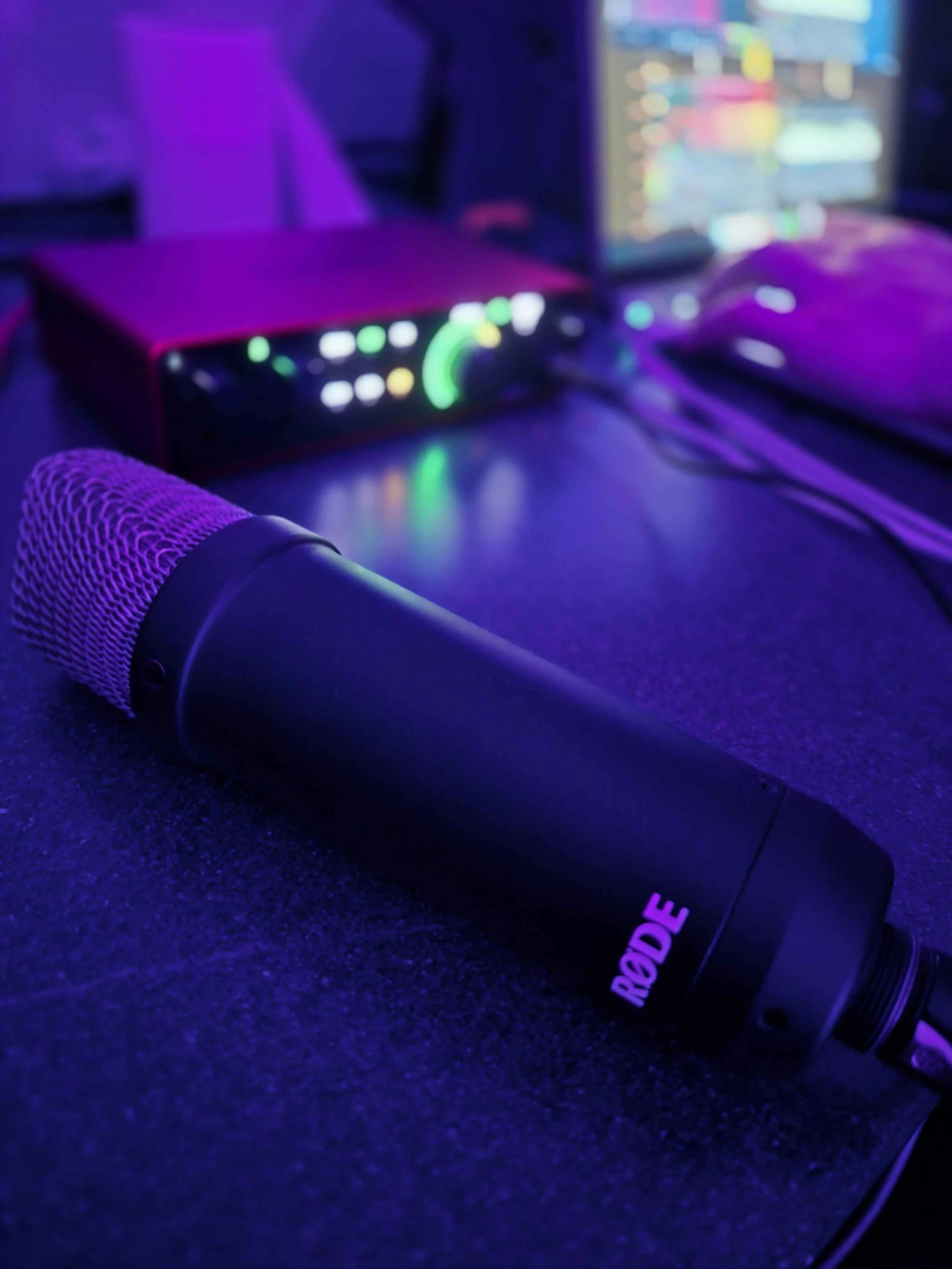 Black microphone with purple lighting, placed on a dark surface with audio equipment and a computer screen in the background.