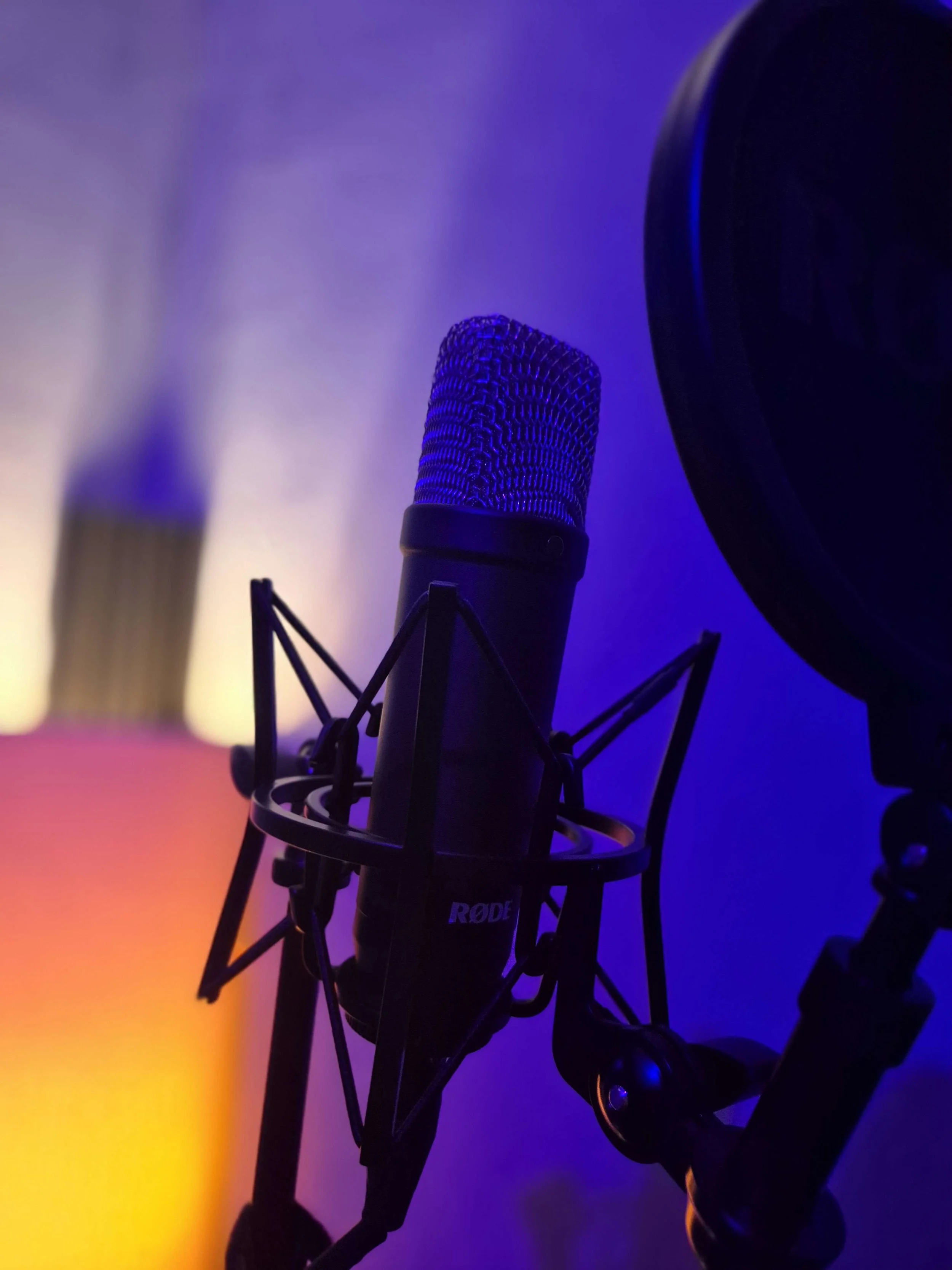 Close-up of a Rode microphone with a pop filter, in a recording studio with colorful background lighting.