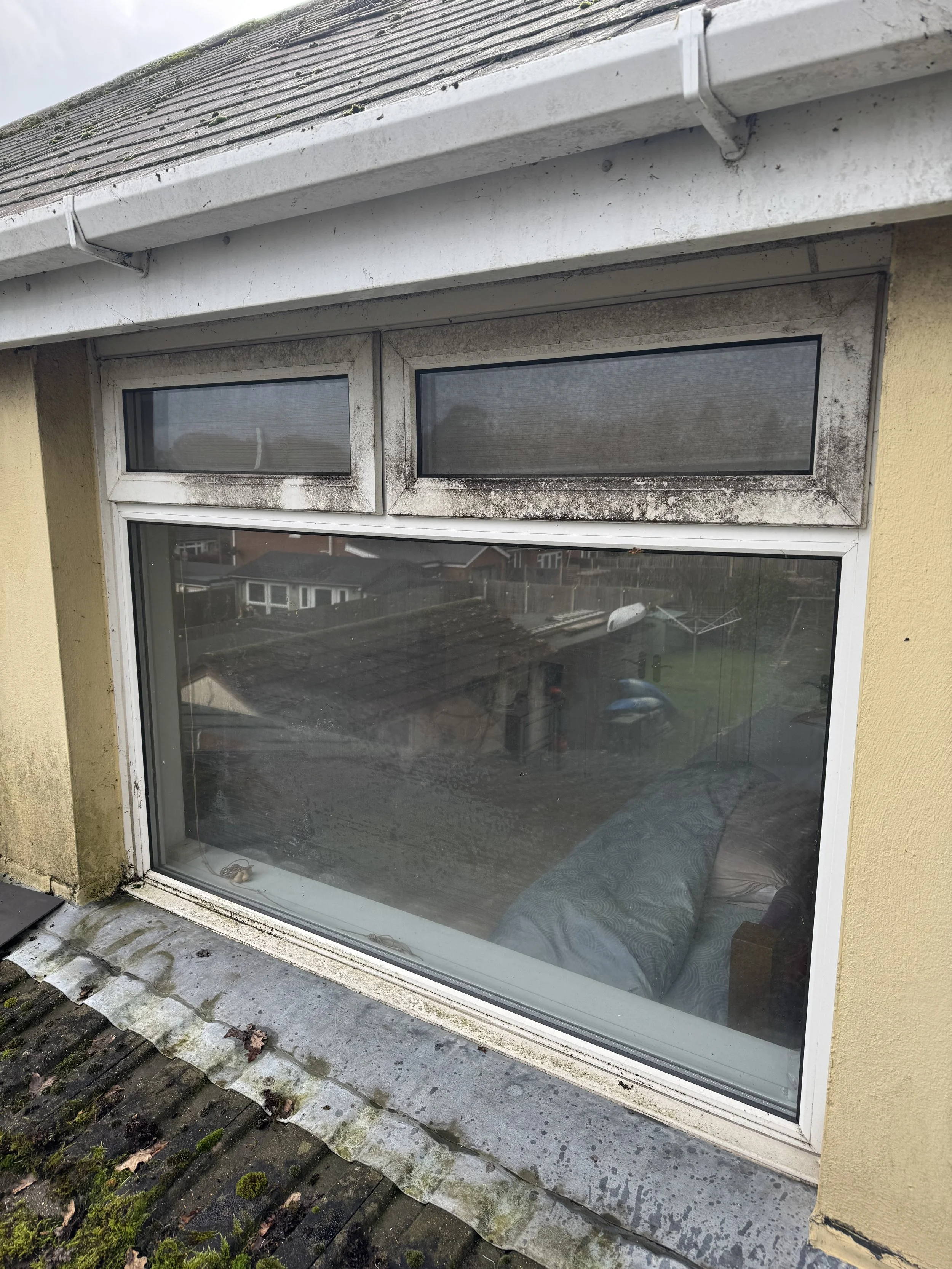 Close-up view of a window with dirty and moldy grime on the window frame, situated in a house with an overhanging roof and mossy, weathered roof tiles outside.