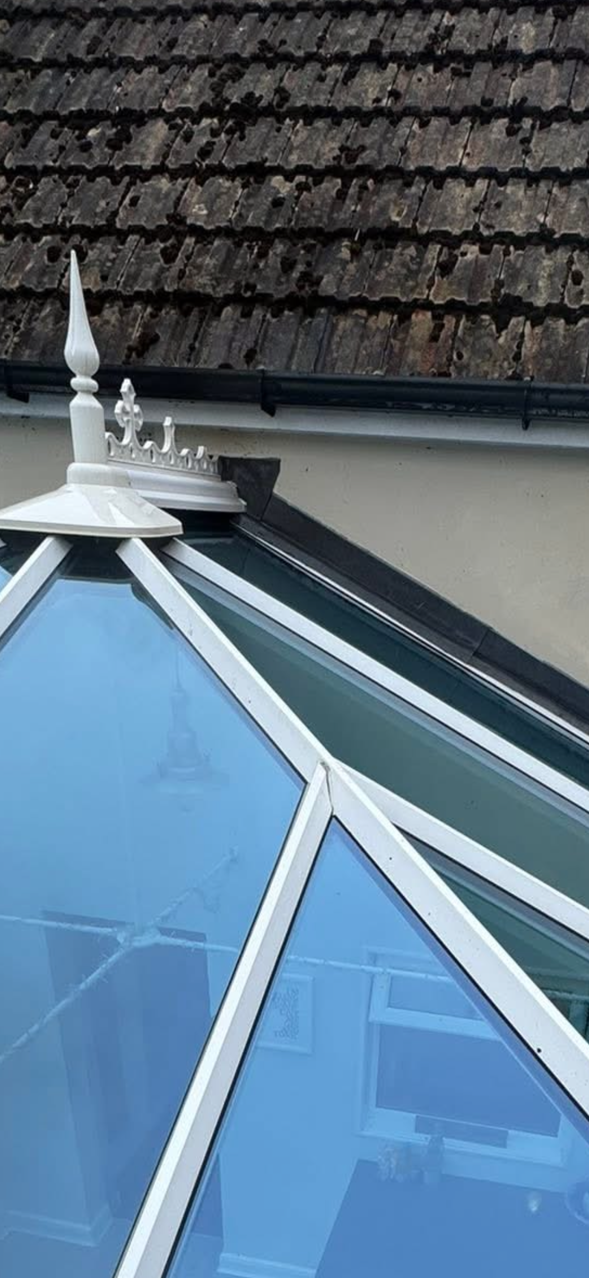 A close-up of a roof with weathered, wooden shingles and a small, white decorative turret on a house. Part of a window with a white frame and triangular glass panes is visible in the foreground.