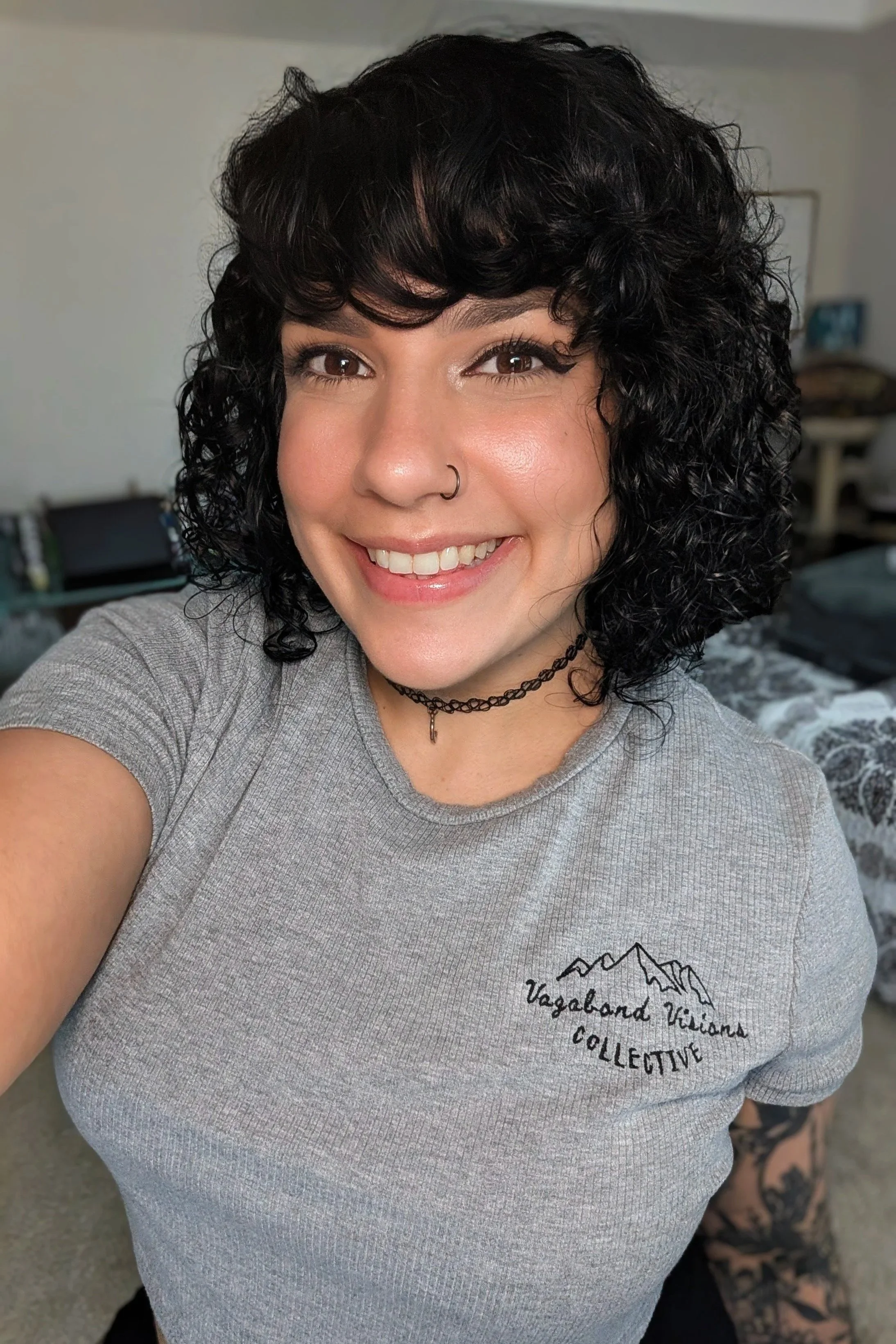 Close-up of a young woman with curly black hair, smiling, wearing a nose ring, a choker necklace, and a gray T-shirt with a mountain and text logo, in an indoor setting.
