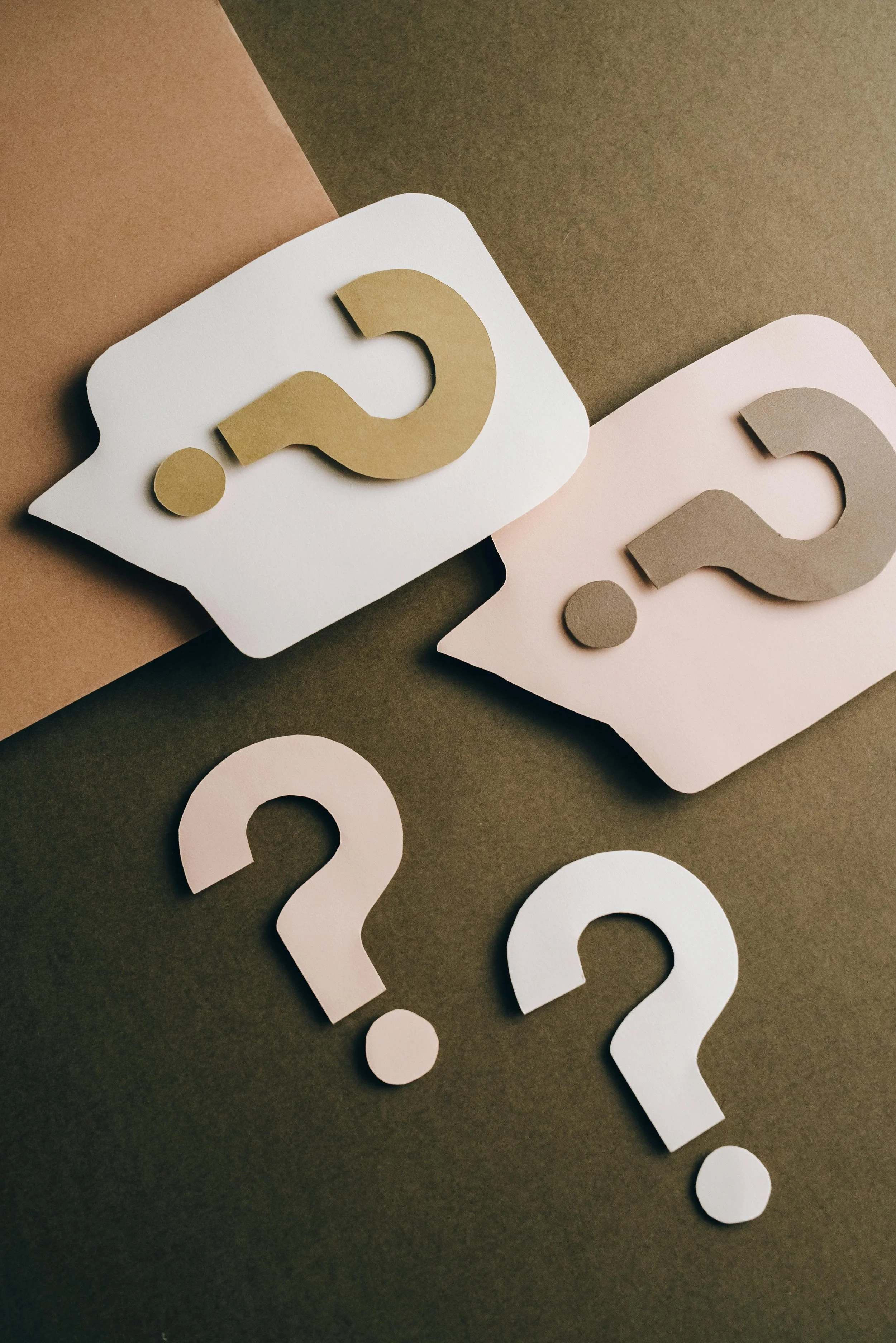 Several paper question marks in white, gold, gray, and pink, scattered on a dark and light background.