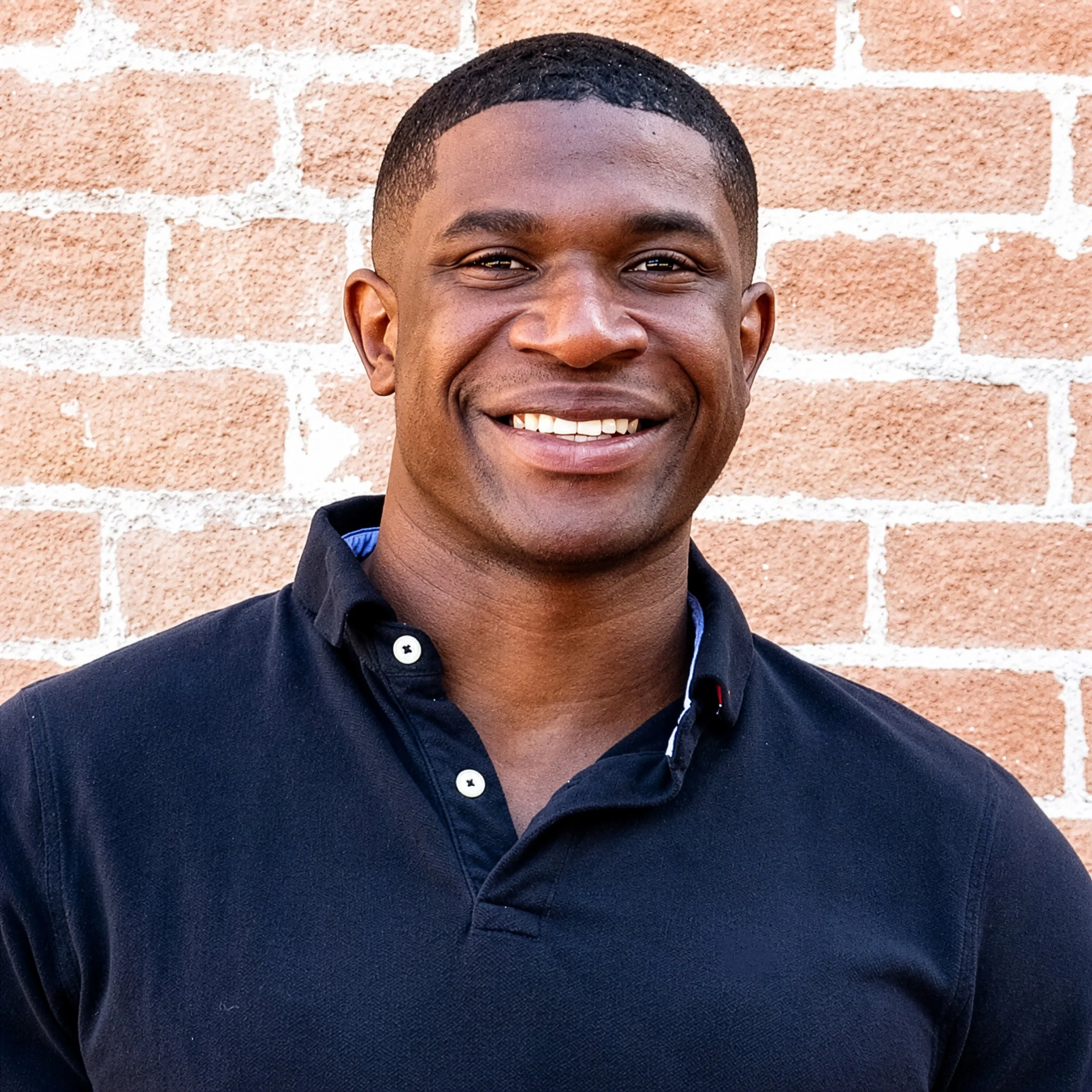 Close-up of a young man smiling with a brick wall in the background.