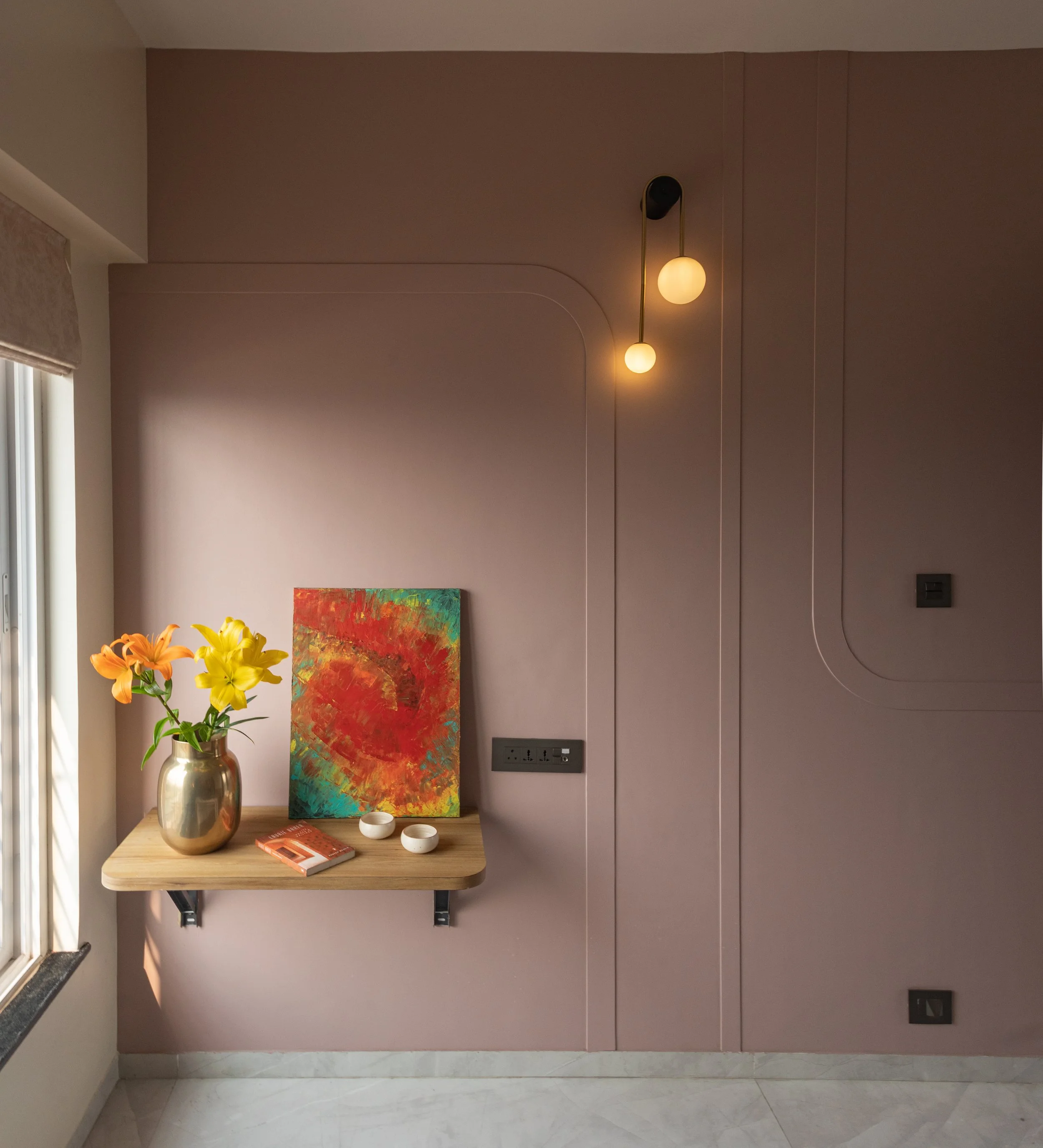 A wooden table with a metallic vase with orange and yellow lilies, an abstract colorful painting, a red book, and two small white bowls, mounted on a mauve wall with built-in electrical outlets and a modern hanging light fixture.