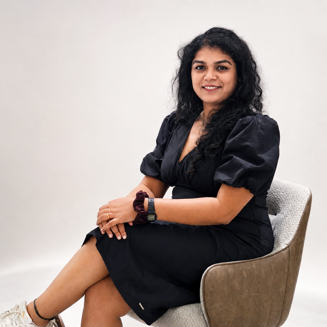 A woman with long curly black hair and medium skin tone sitting on a modern beige chair. She is wearing a black dress with puffed sleeves and white sneakers with black ankle socks, sitting against a plain white background. She is smiling and looking at the camera.
