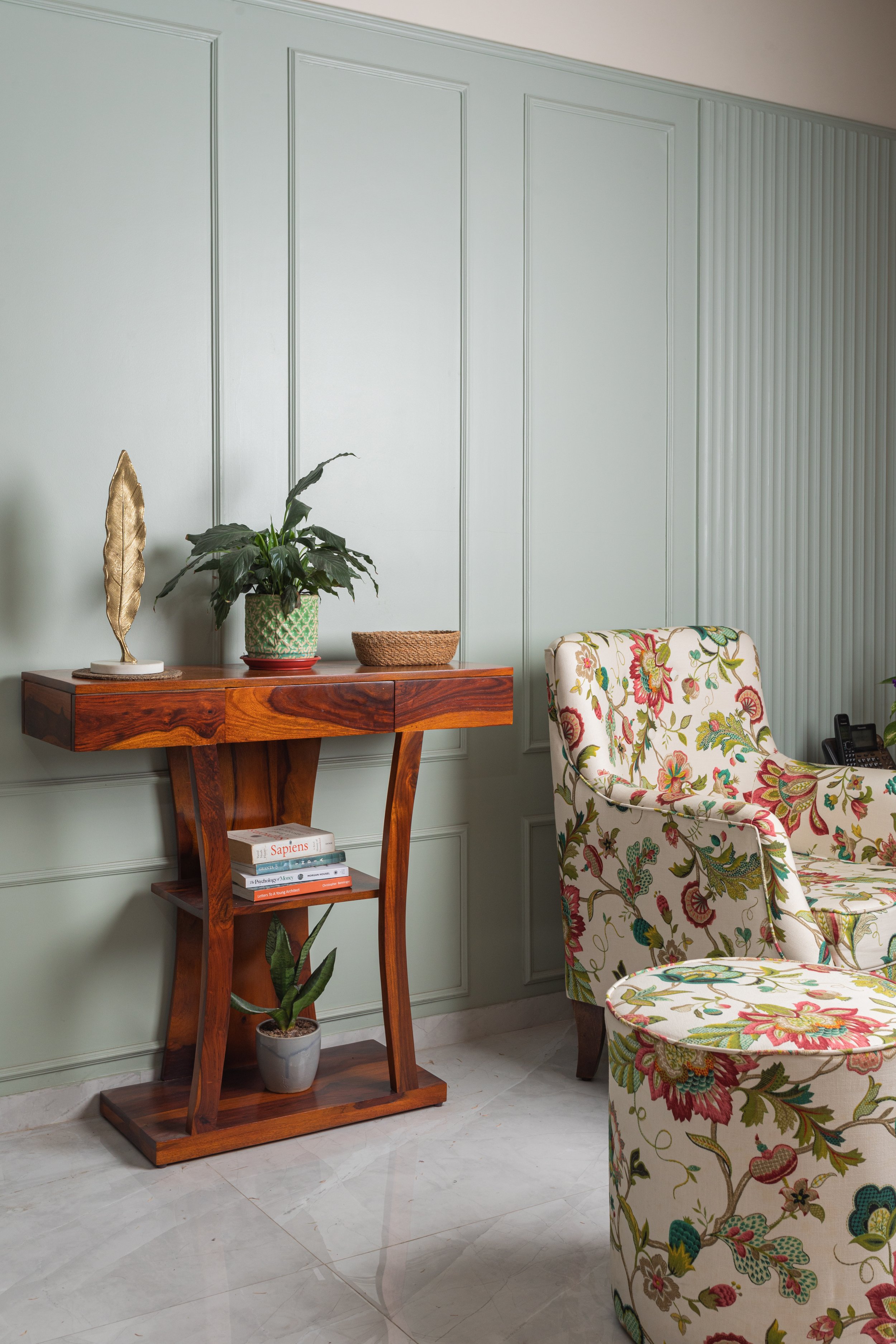 Interior with light green paneled walls, wooden console table holding a decorative gold feather sculpture, green plant in a patterned pot, and a woven basket. A floral upholstered armchair is next to the table, and a potted plant is in the corner.
