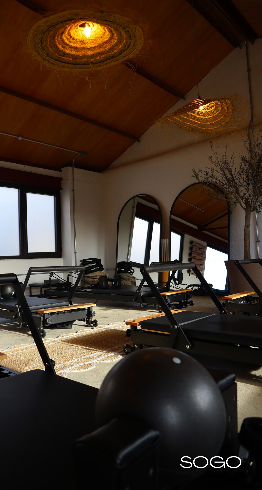 A Pilates studio with a wooden ceiling, two decorative woven pendant lights, large windows with frosted glass, mirrors, and Pilates reformer machines arranged on a rug. There is a small tree in the corner and a logo that reads 'SOGO' in the bottom right corner.