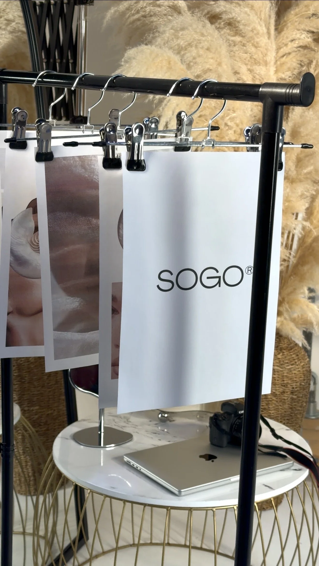 A clothing rack with SOGO branded papers clipped to it, situated in a room with a marble-top table holding a closed laptop, a camera, and a mirror, with decorative pampas grass in the background.