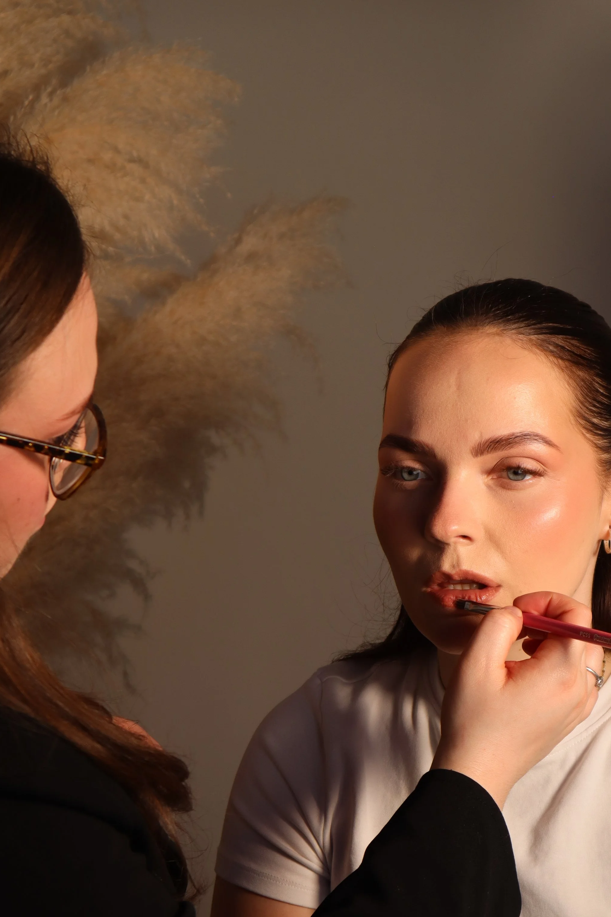Makeup artist applying lipstick to a woman with dark hair, wearing a white t-shirt, against a gray background.