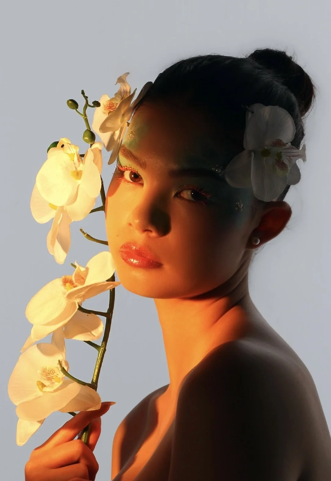 A woman with dark hair and makeup holding a stem of white orchids, with the orchids partly covering her face, and illuminated by warm, golden sunlight.