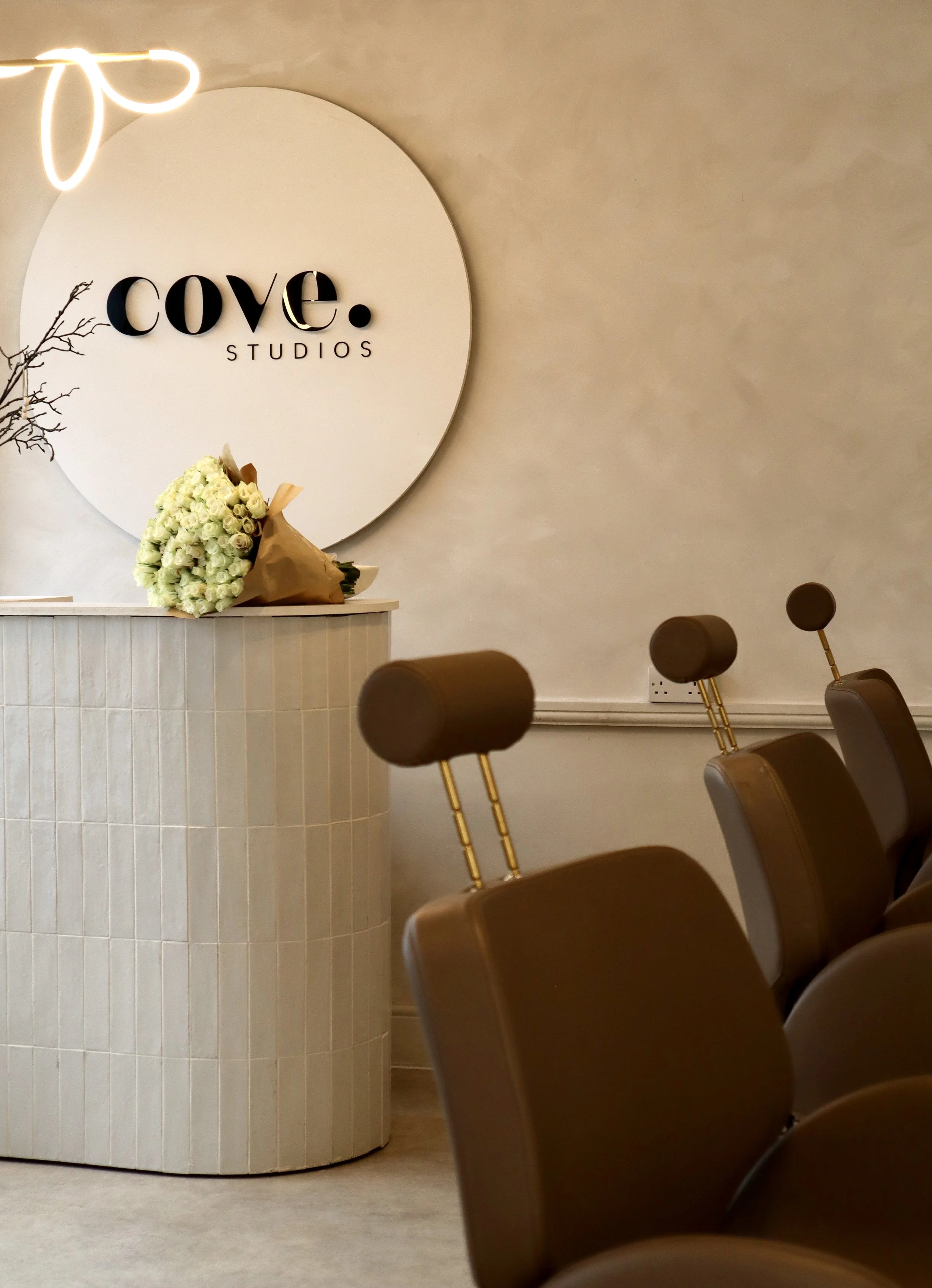 Interior of a studio reception area with a white round sign that reads 'cove STUDIOS', a bouquet of white flowers on a curved counter, and brown chairs with headrests.