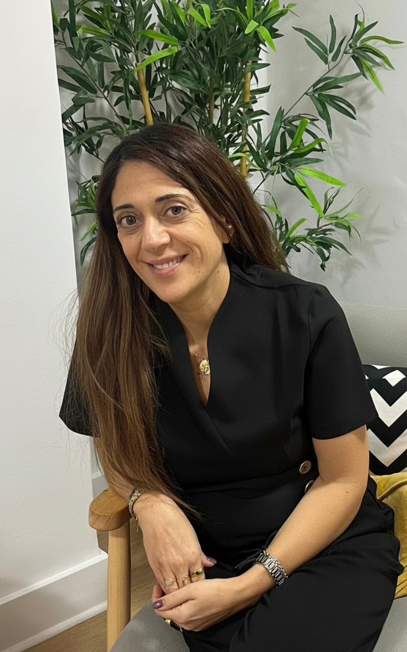 A woman with long brown hair sitting on a beige chair in front of a large green plant. She is smiling and wearing a black outfit with jewelry, including rings, a watch, and a necklace. There is a black and white chevron-patterned pillow behind her.
