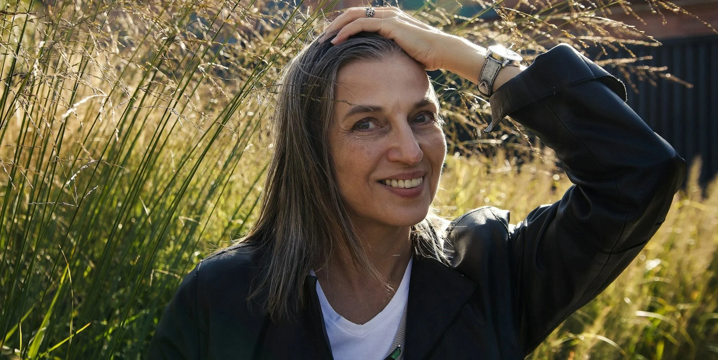 A woman with long brown hair and a black leather jacket smiling outdoors during the daytime, with her hand on her head, standing in front of tall golden grass.