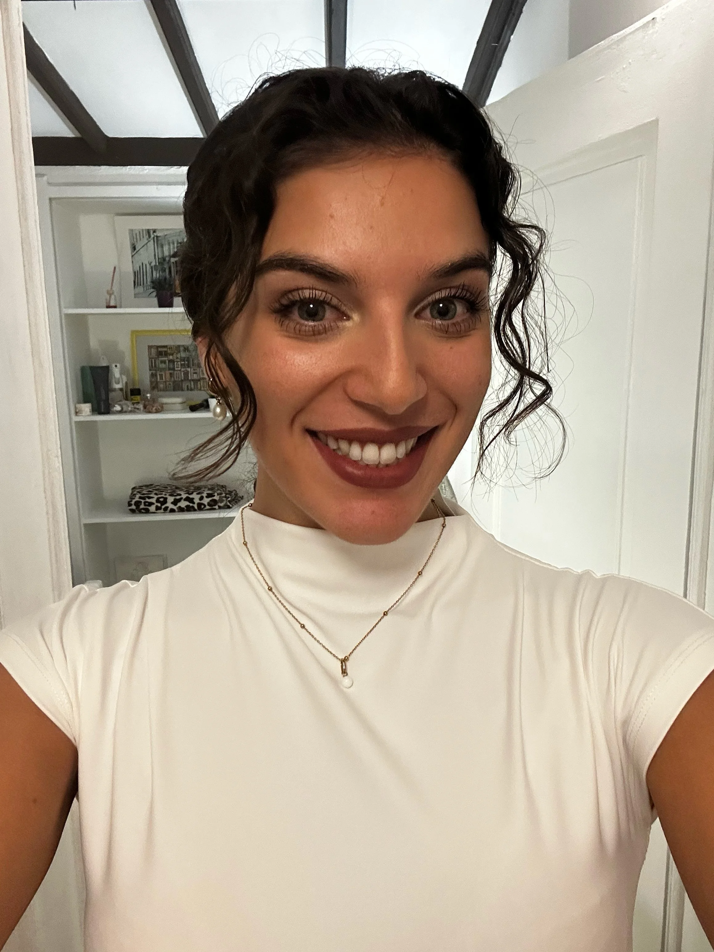 Close-up of a smiling woman with curly dark hair, wearing a white shirt and layered gold necklaces, in a room with white walls and a shelf in the background.