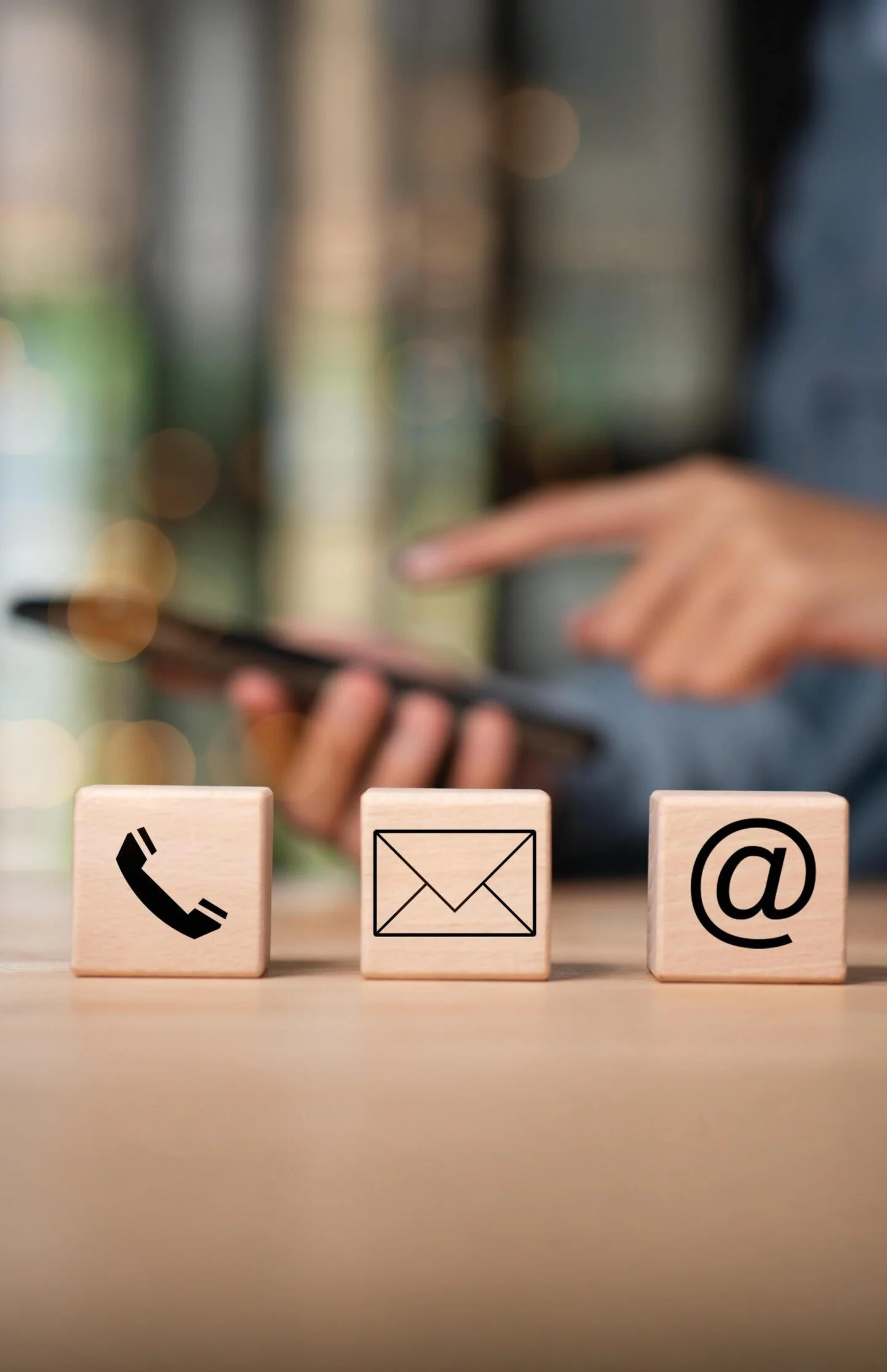 Three wooden blocks with icons of a phone receiver, an envelope, and an at symbol (@) on a table. In the blurred background, a person is holding a smartphone.