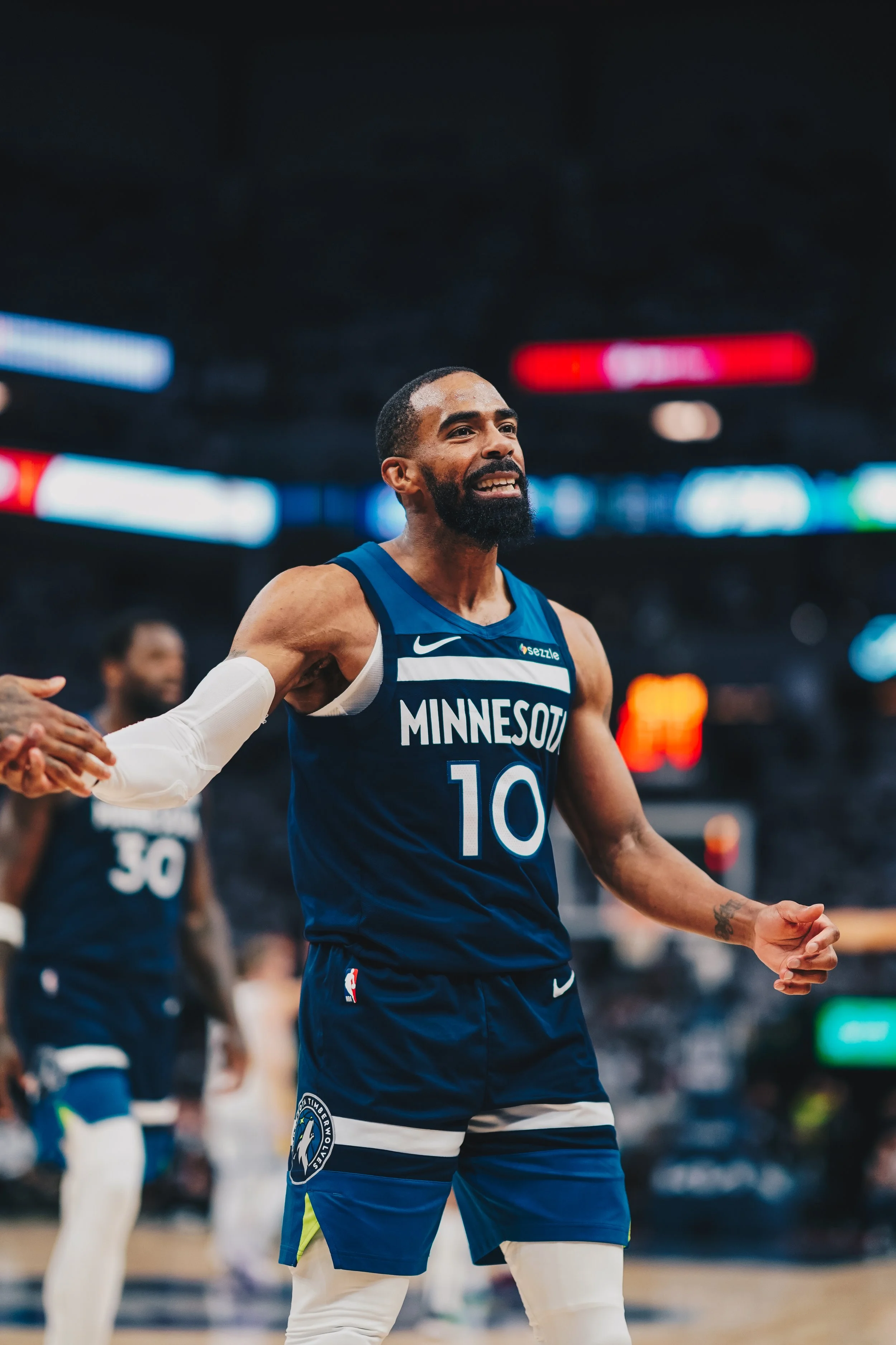 An African American male basketball player wearing a blue Minnesota Timberwolves jersey with the number 10 is smiling and holding hands with a teammate on the basketball court during a game.