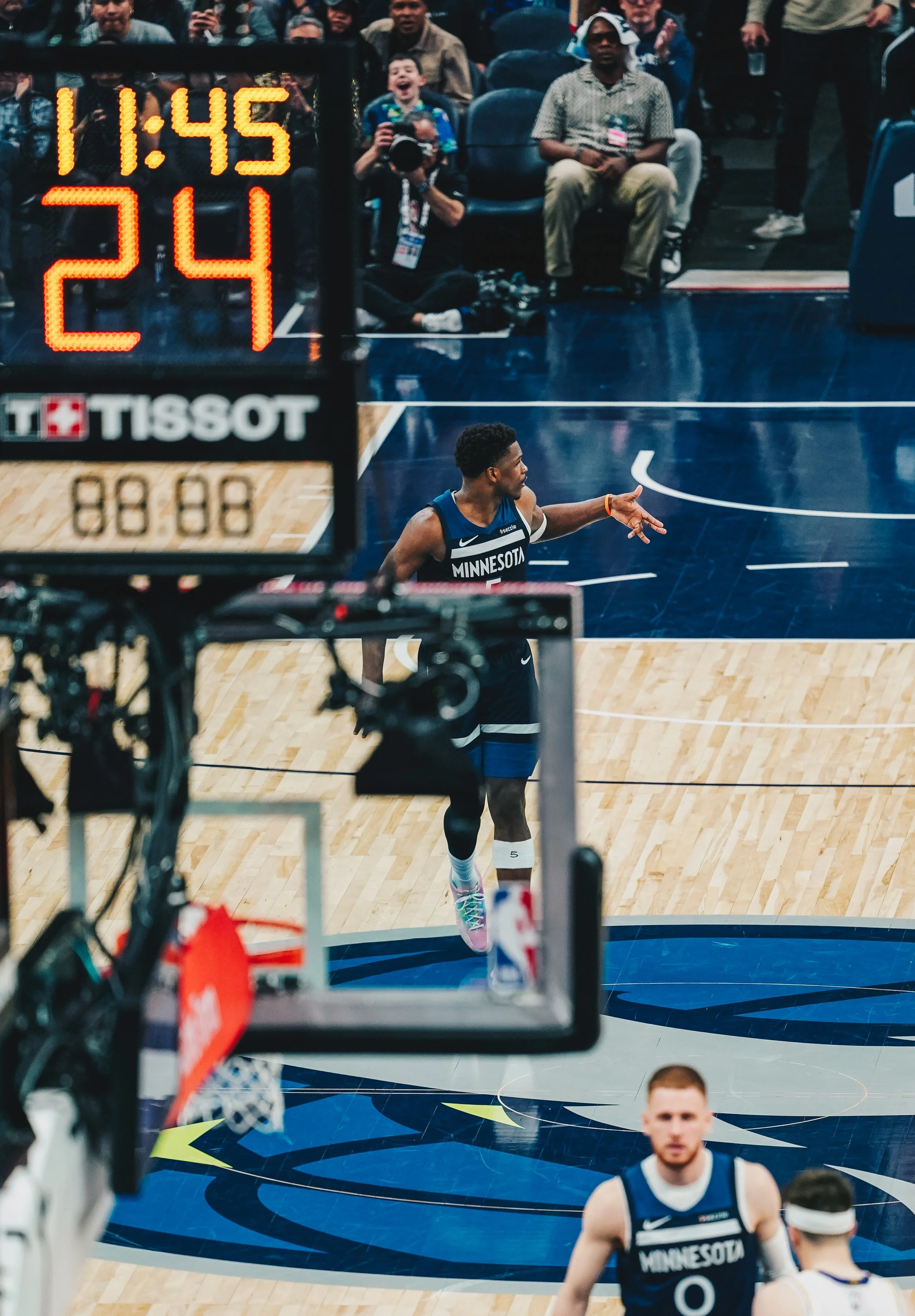An NBA player in a Minnesota Timberwolves uniform pointing on the court during a game, with players and spectators visible in the background.