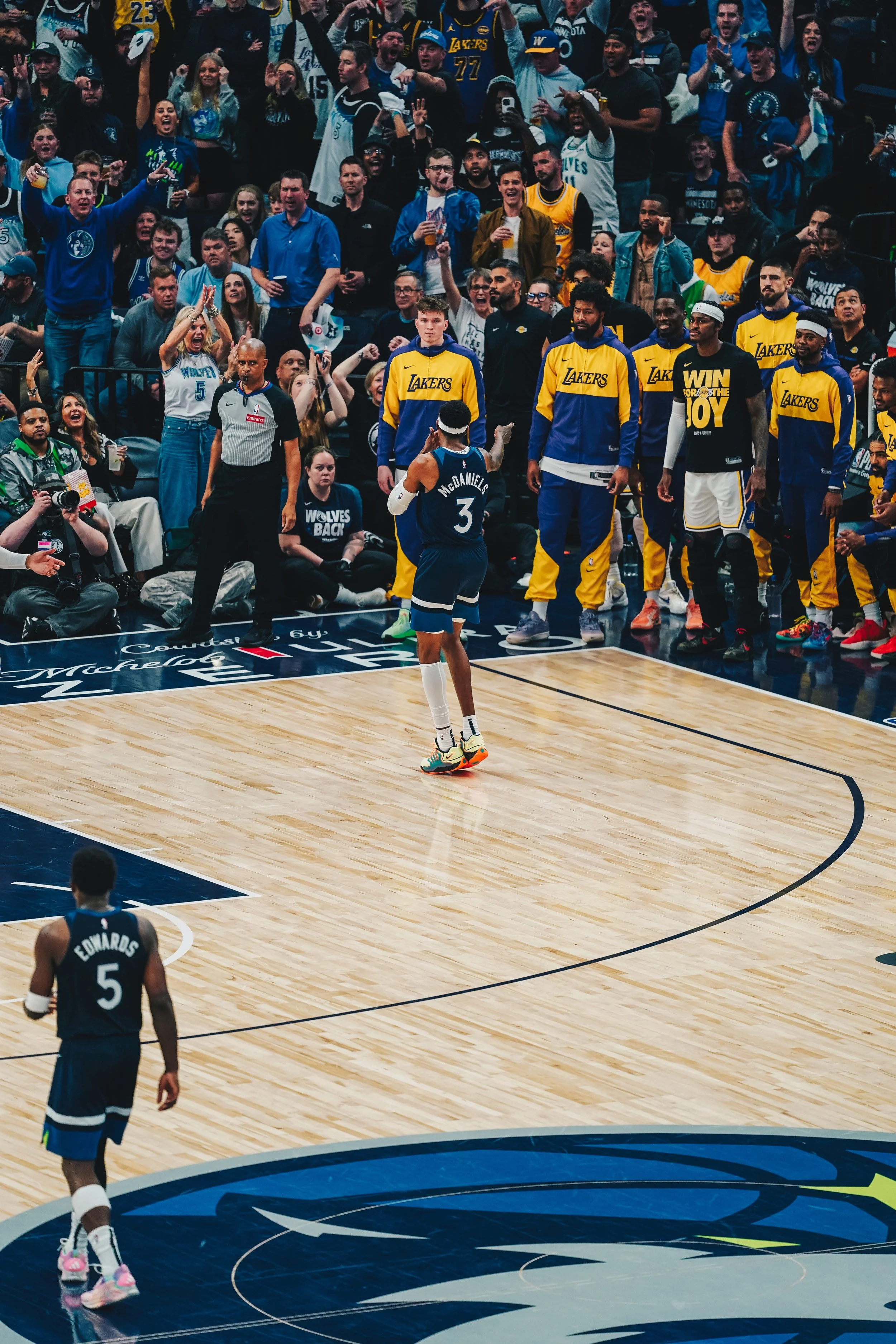 Basketball player in navy uniform with the name 'McDaniels' on the back, standing on a basketball court with crowd of fans in the background, some wearing Lakers and Wolves jerseys, cheering and taking photos.