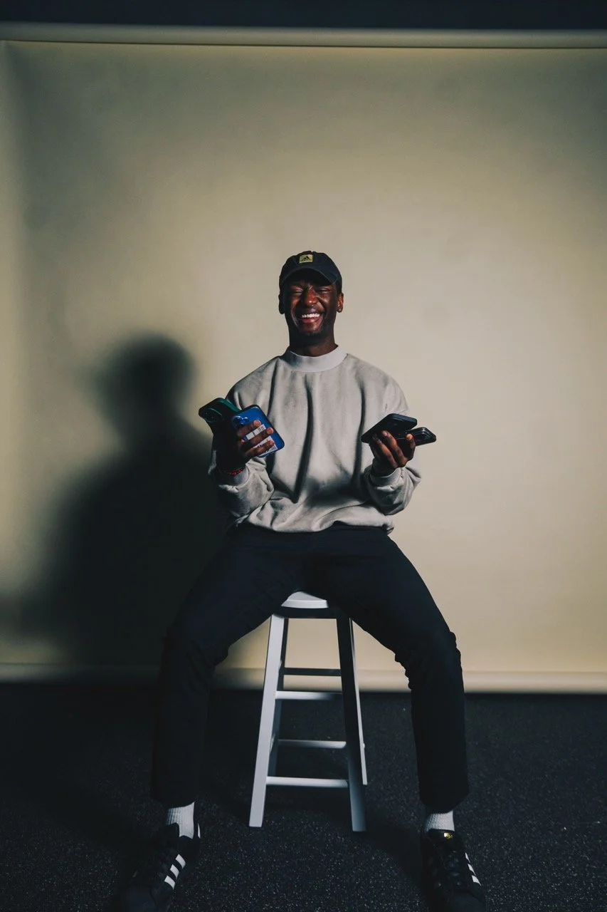 A young man sitting on a white stool, holding multiple phones in his hands, smiling and laughing while wearing a gray sweatshirt, black pants, black sneakers, and a black cap, in front of a plain beige wall.