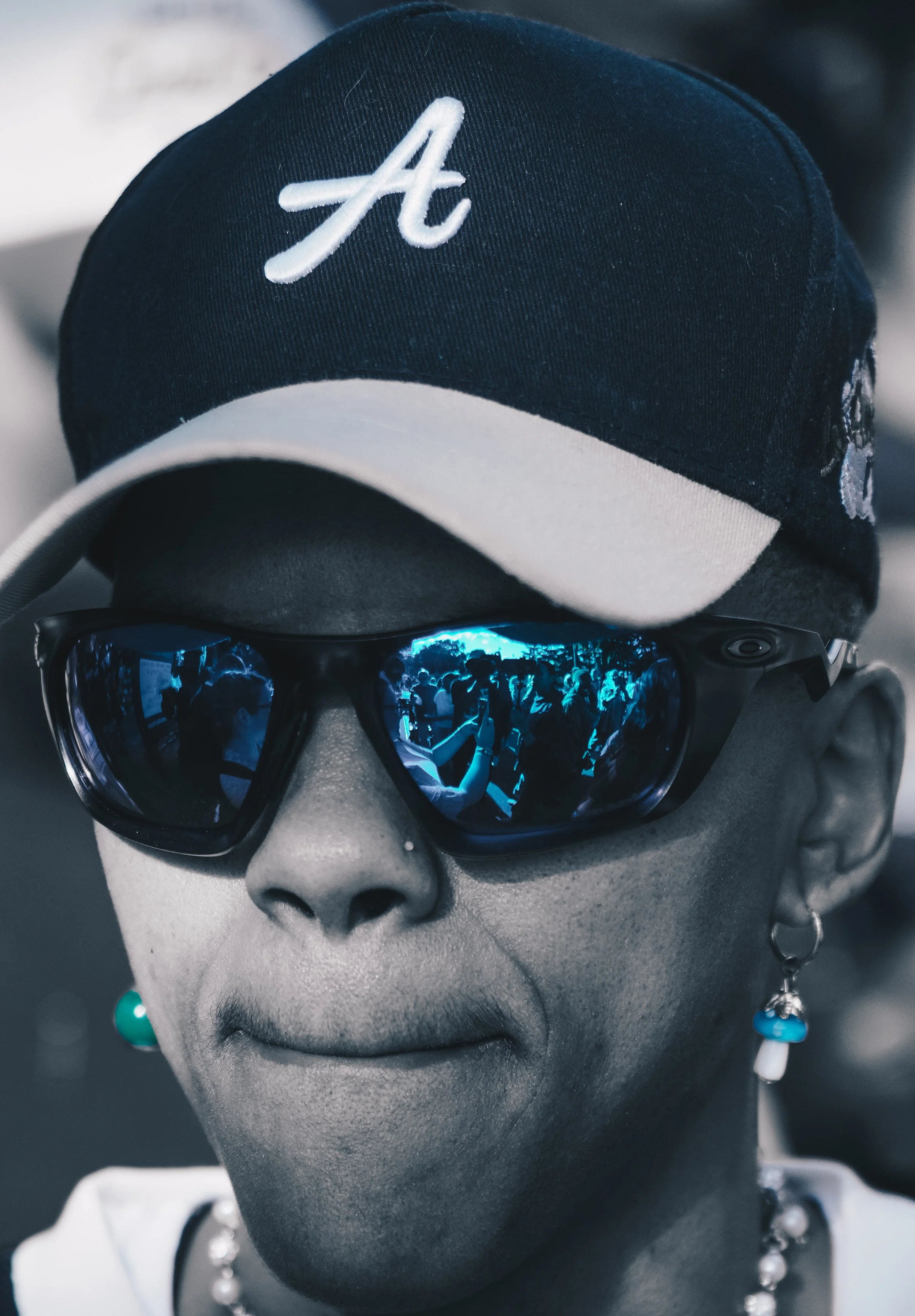 A mannequin wearing a black and white baseball cap with an embroidered white letter A, sunglasses with reflective blue lenses showing a crowd of people, and earrings, with a blurred background.