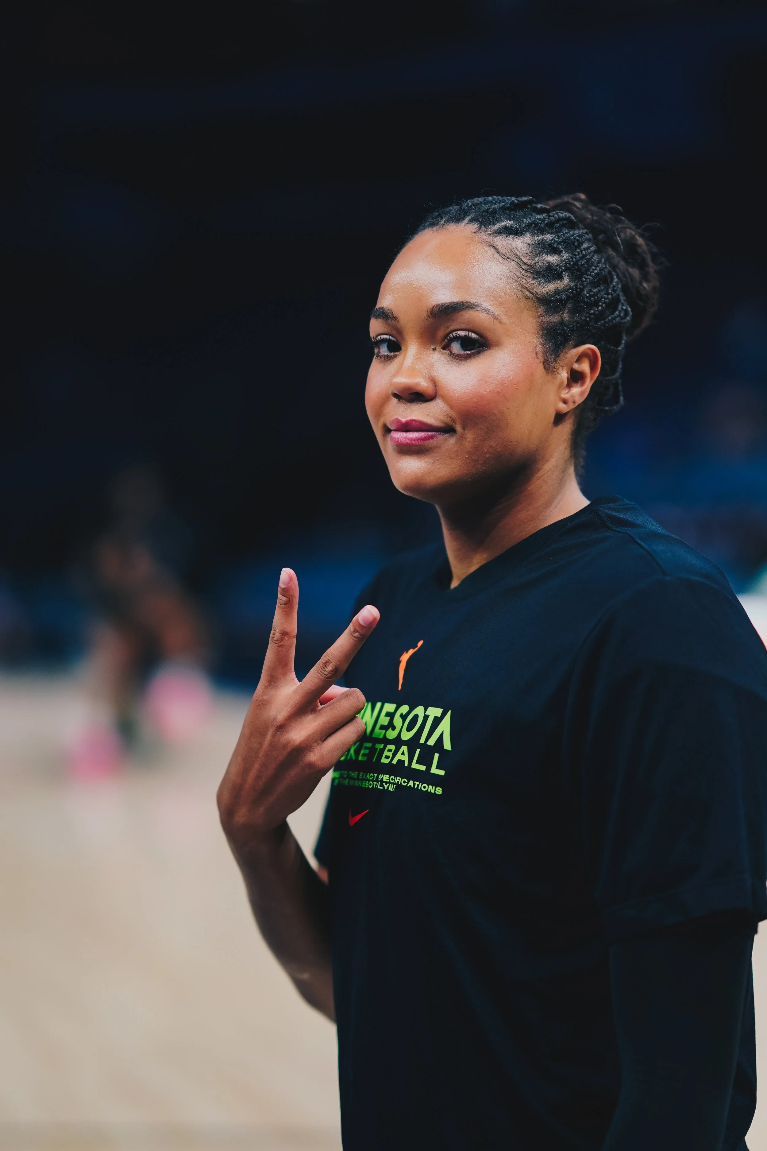 A young woman with braided hair, wearing a black Minnesota basketball t-shirt, making a peace sign gesture and looking at the camera in an indoor sports arena.