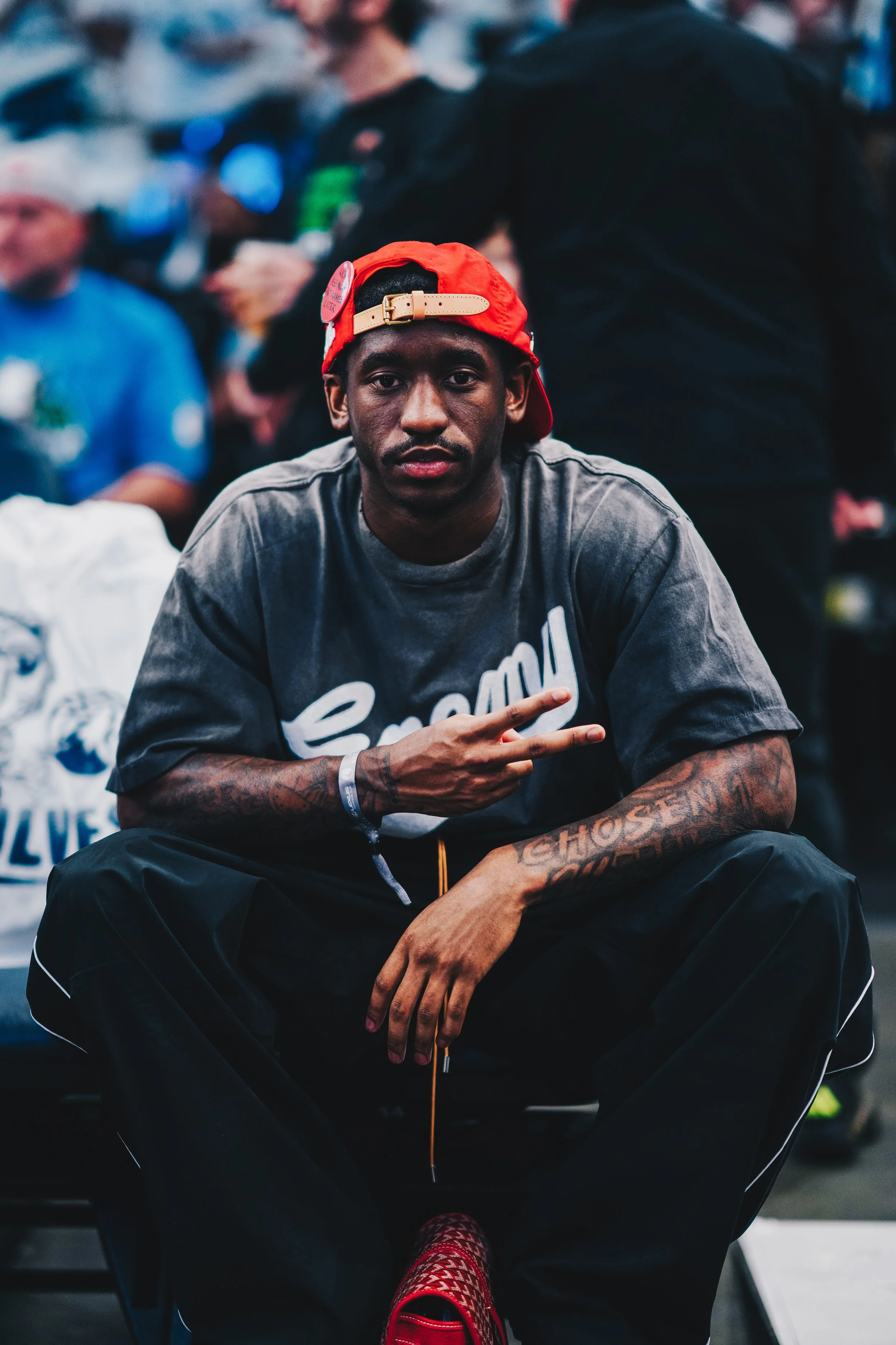 A young man with dark skin wearing a red backwards cap, a grey t-shirt, and black pants, sitting with his legs apart and making a peace sign with his right hand, in a busy, crowded setting.