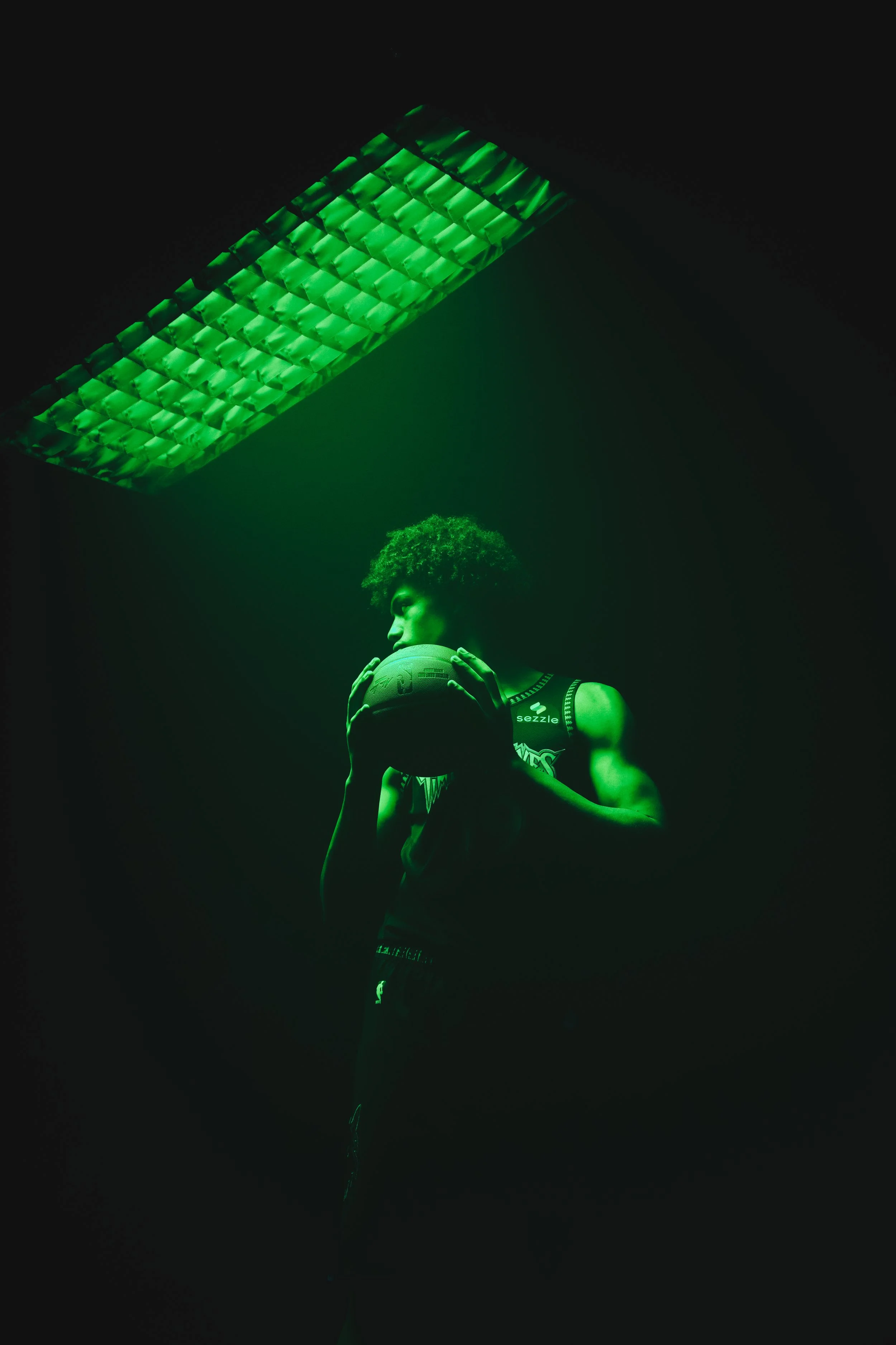 A basketball player in a dark room with green lighting, holding a basketball and looking down.