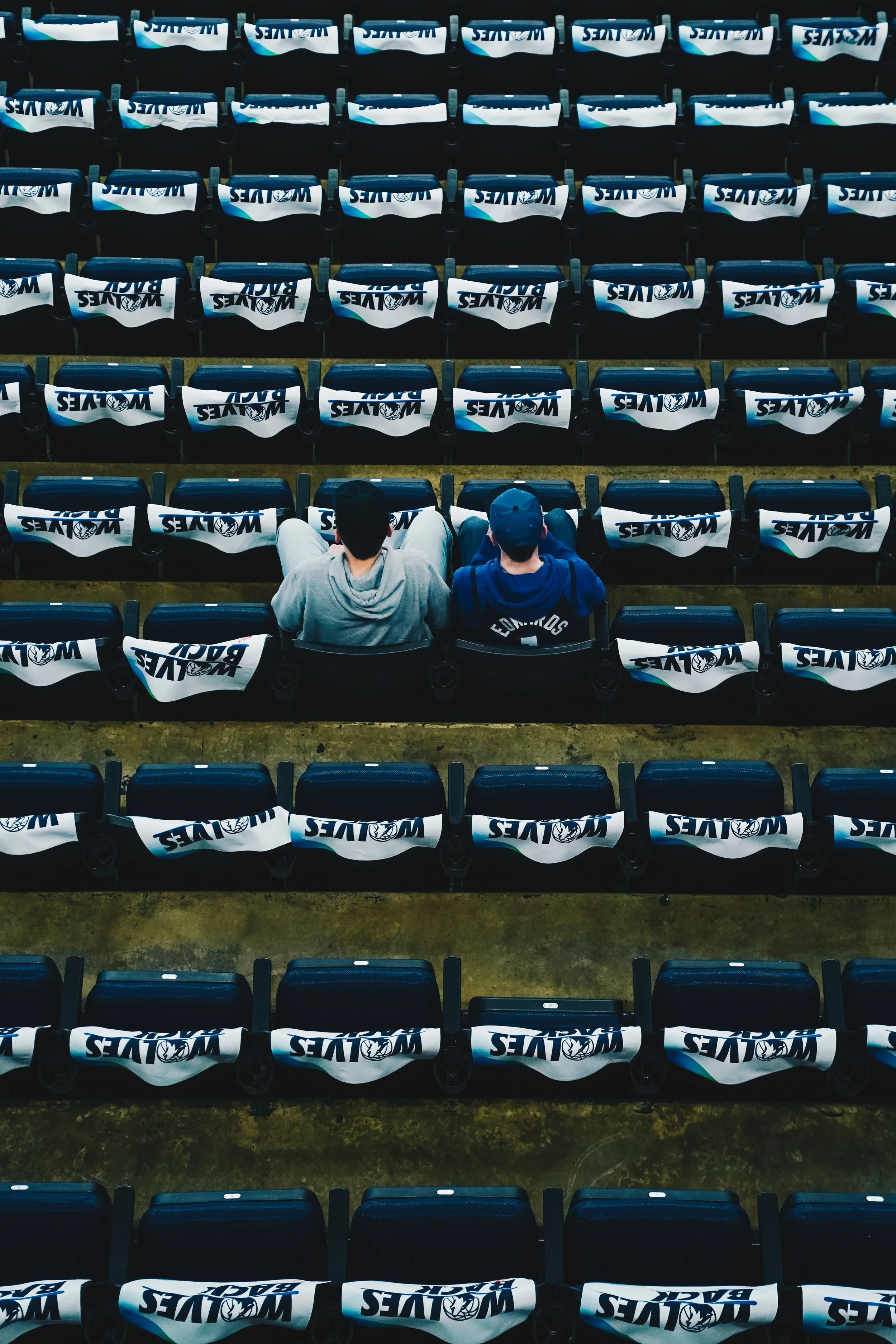 Two people sitting in empty stadium seats with WOLFTEAMS logo on the seat backs, viewed from behind.