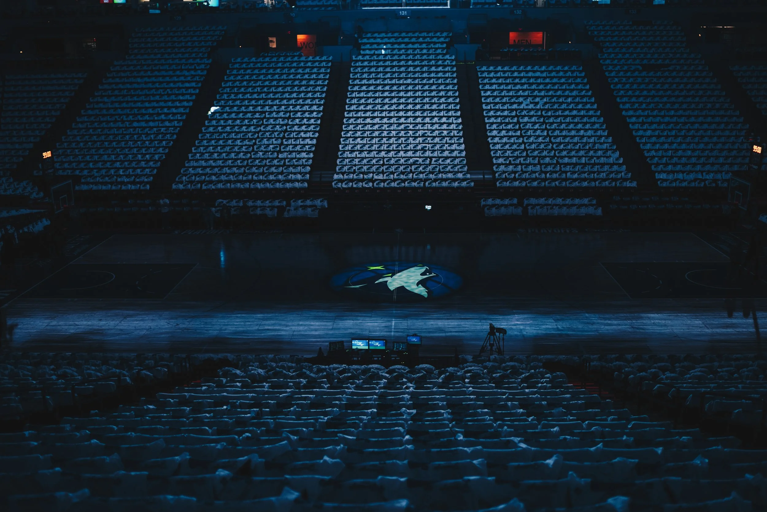 Empty basketball arena with blue seats, illuminated by arena lights, featuring a basketball logo on the court.