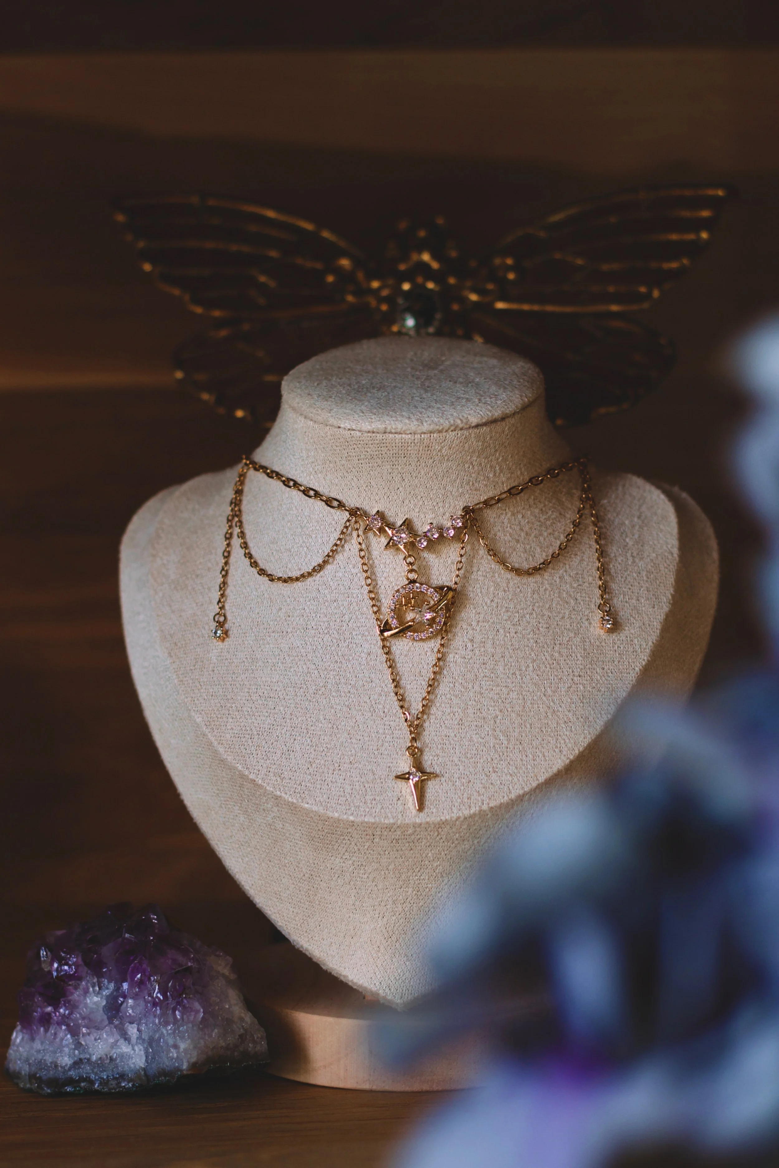 Collier en chaîne dorée avec pendentifs étoiles et cristaux, présenté sur un buste en tissu beige, avec une décoration avec une aile en métal doré en fond et une améthyste en pierre naturelle en bas à gauche.
