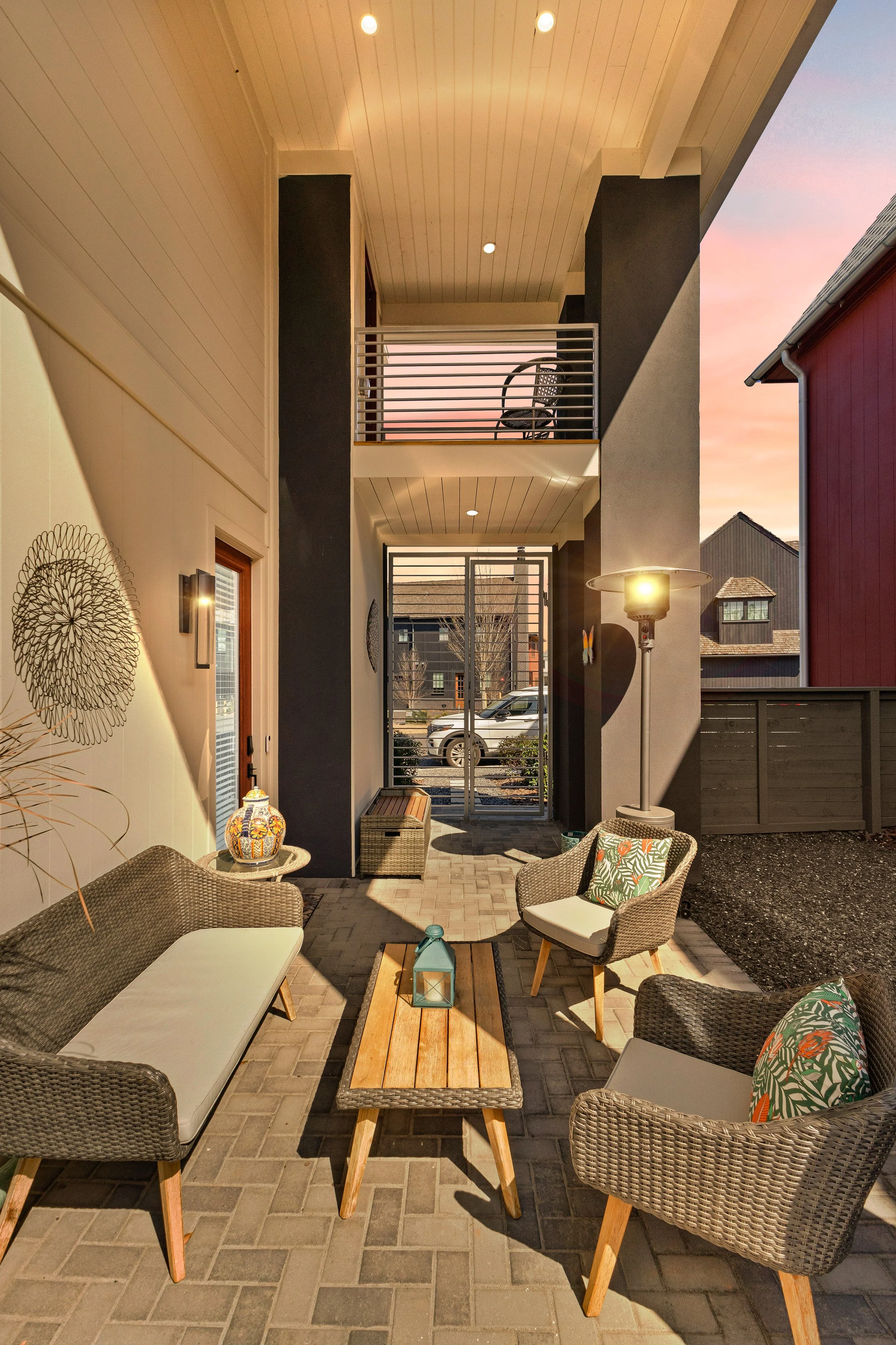 Outdoor patio with wicker chair and bench, wooden coffee table with lantern, decorative pillows, lamp, and potted plant, enclosed by house walls and a gate, at sunset.