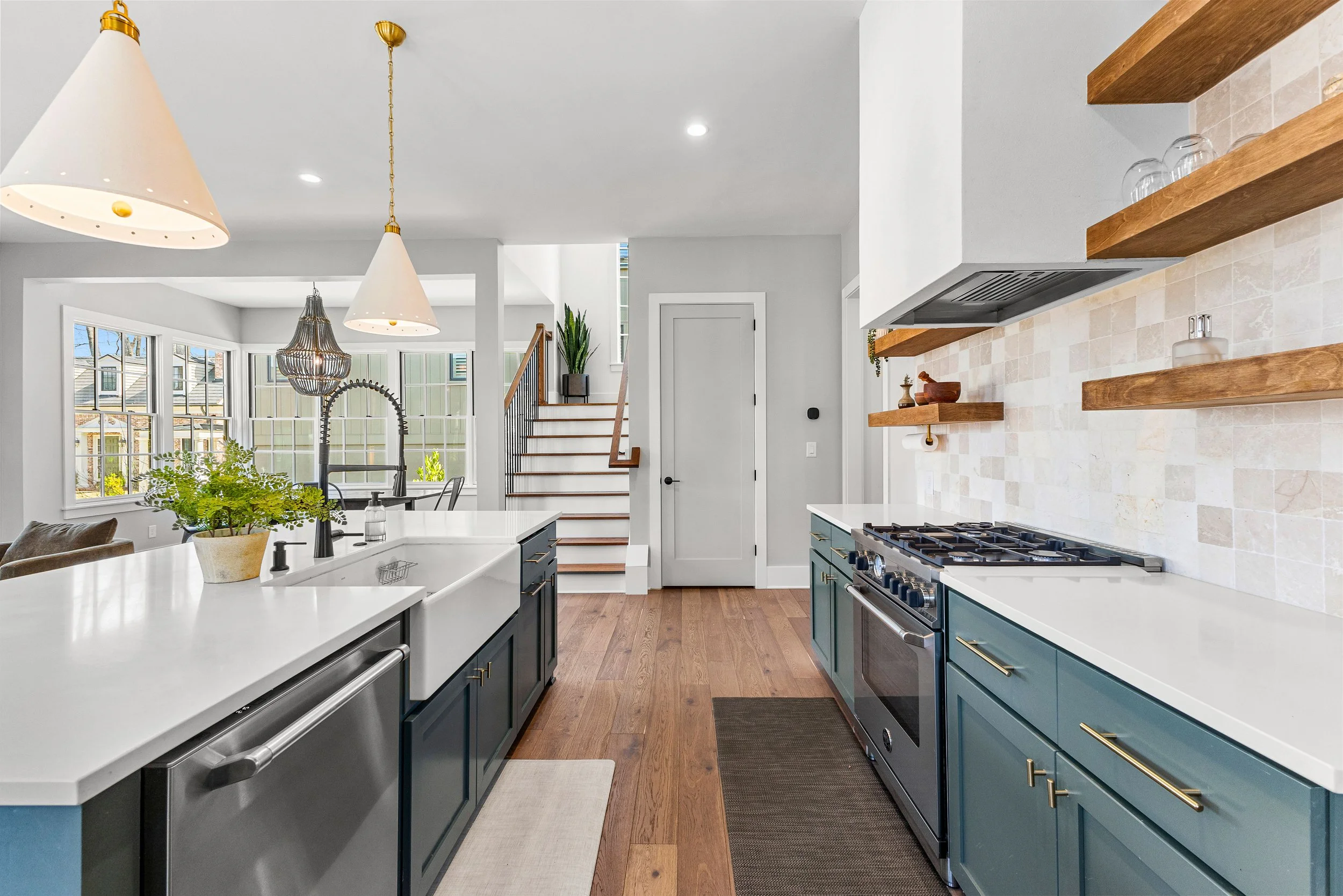 Modern kitchen with blue cabinets, white countertops, and a white farmhouse sink. Open shelving with glassware, a stainless steel stove, hardwood floors, and a view of a dining area and staircase with plants.