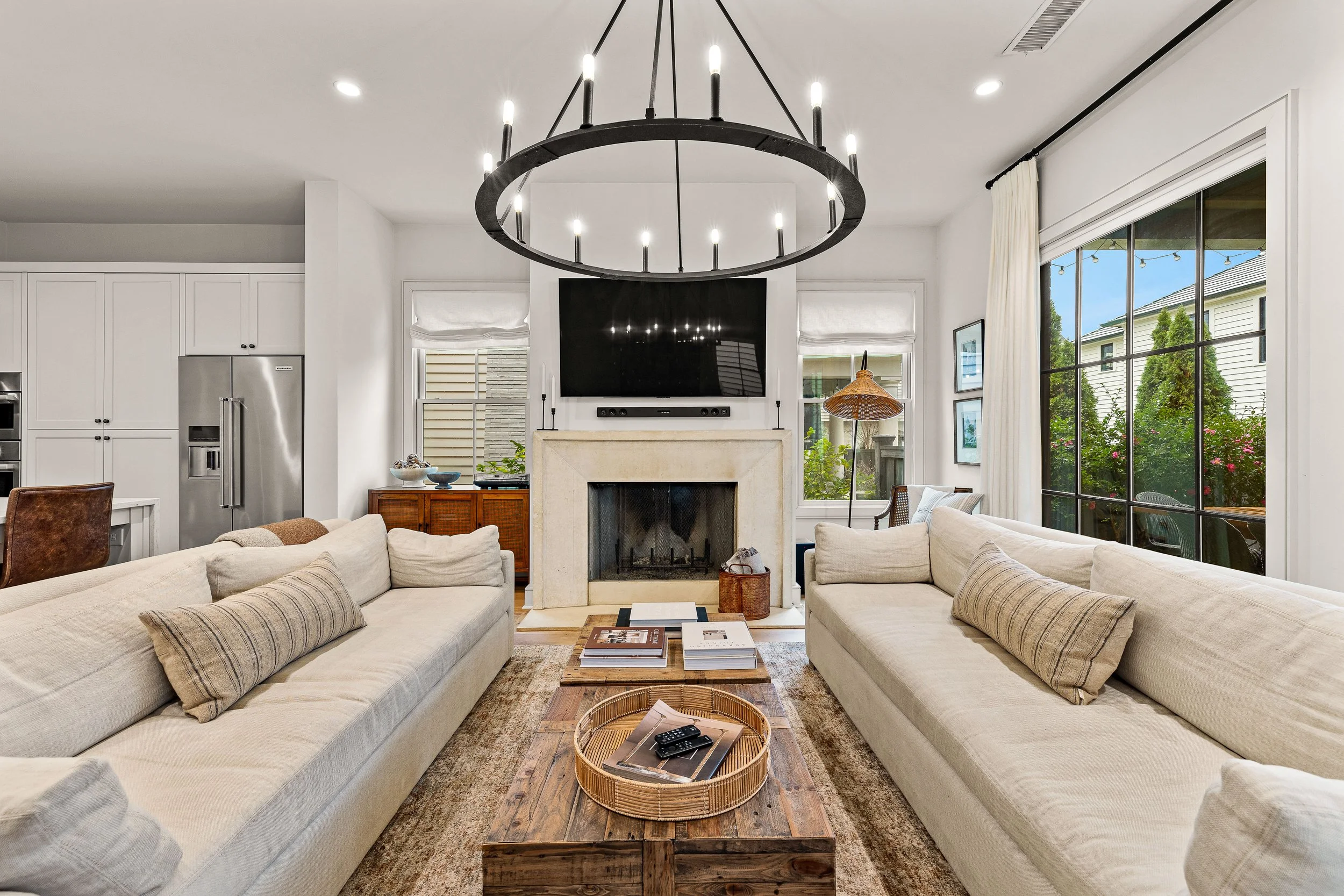 Living room with two cream-colored sofas facing each other, wooden coffee table in the center, fireplace beneath a mounted flat-screen TV, large window with black framing and white curtains to the right, decor and accessories including a round basket, books, and remote control.