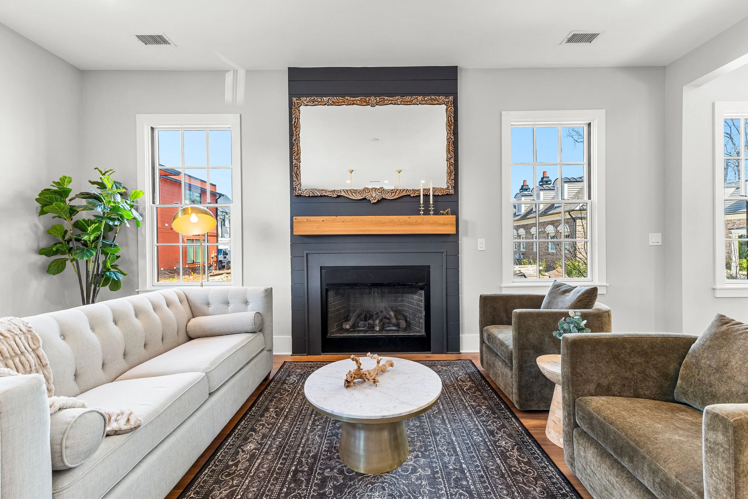 Living room with a fireplace, large mirror above the mantel, and three windows showing a neighborhood outside. Furniture includes a white tufted sofa, two gray armchairs, a round marble coffee table, and a large potted plant.