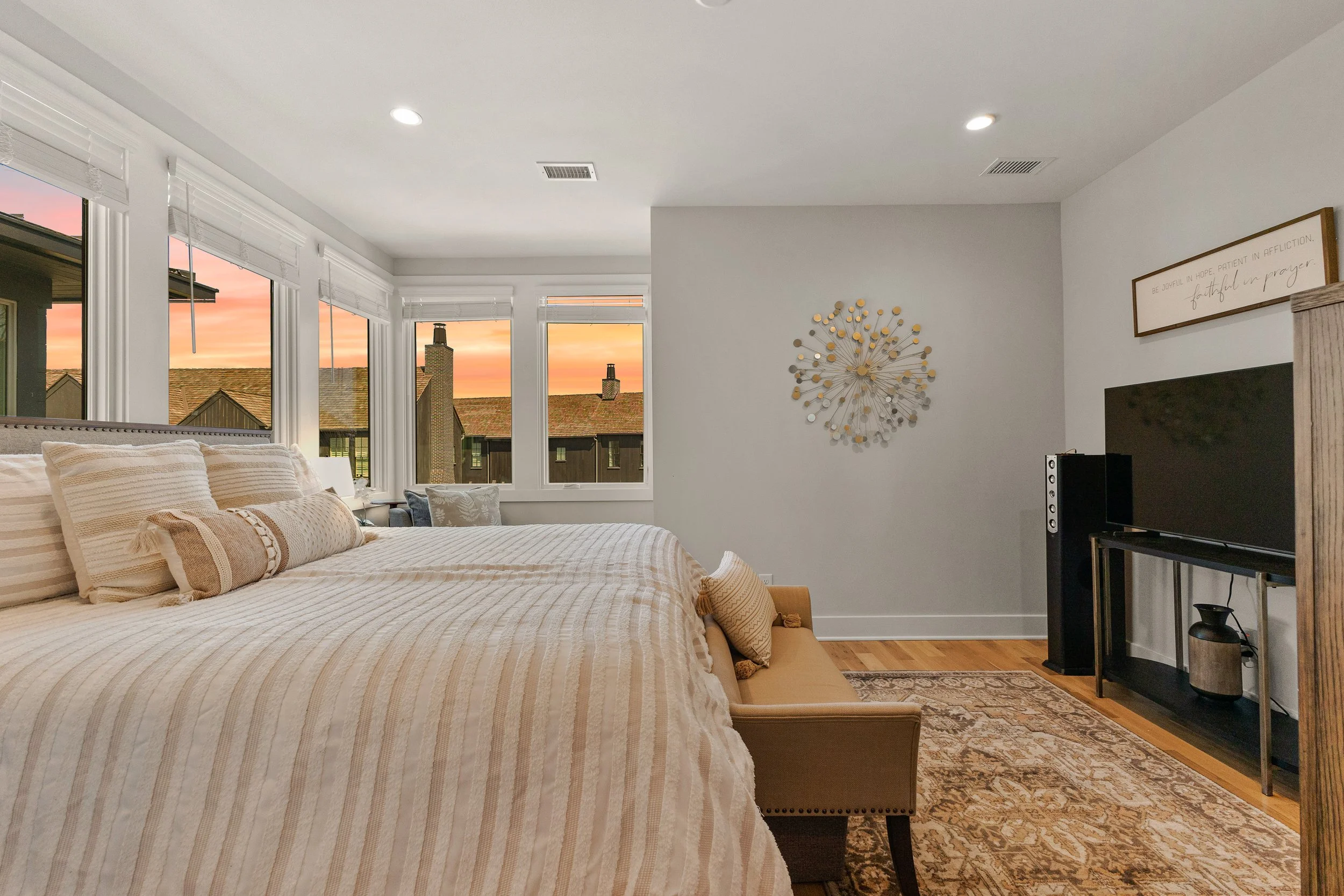 A bedroom with a large bed covered in cream and beige bedding, surrounded by pillows. Multiple windows showing a sunset sky, rooftops, and chimneys outside. Wall decor and a TV on a stand are visible.
