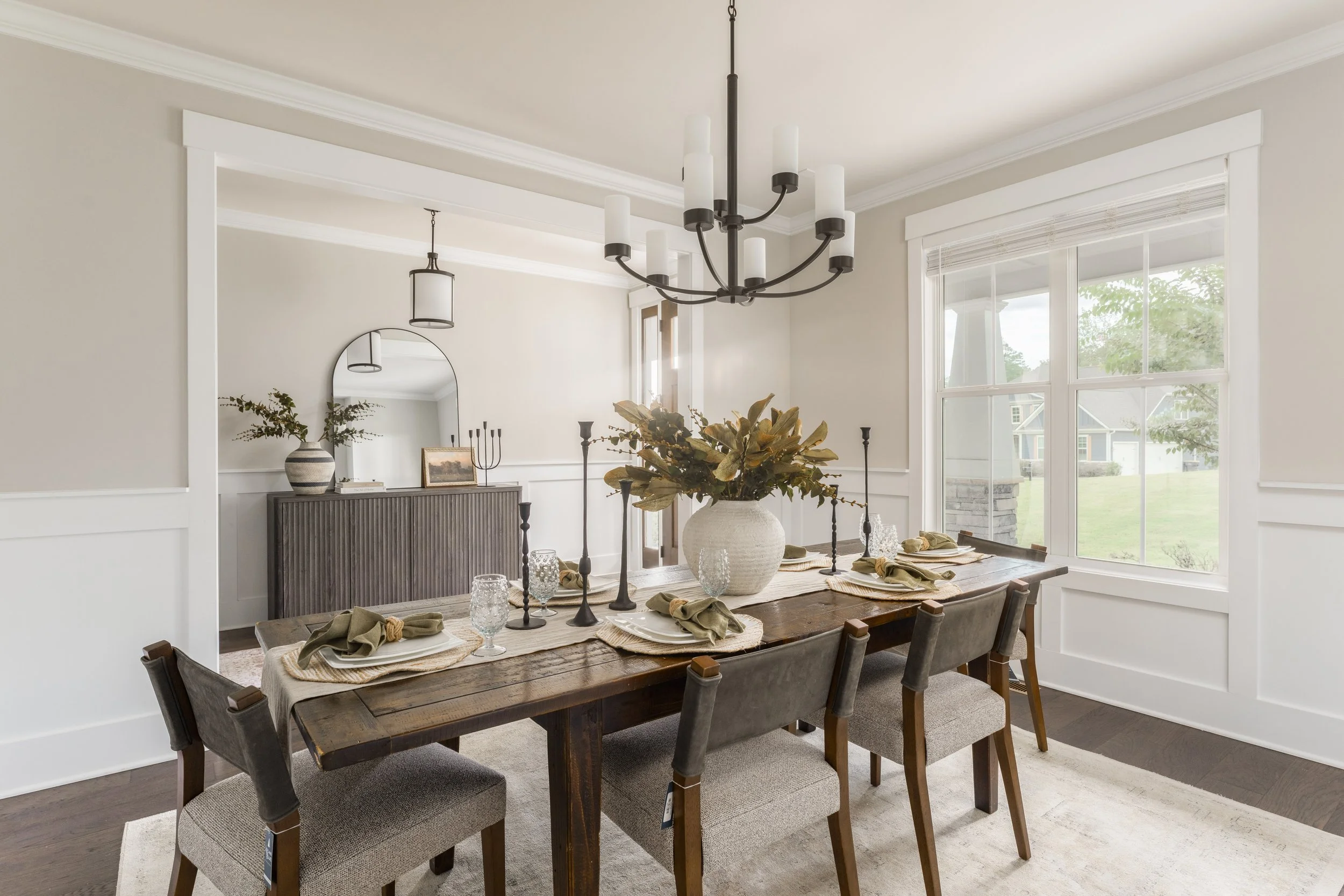 Dining room with wooden table, six chairs, large window, chandelier, vase with greenery, placed on a white runner, beige walls, and a sideboard with mirror and decor items.