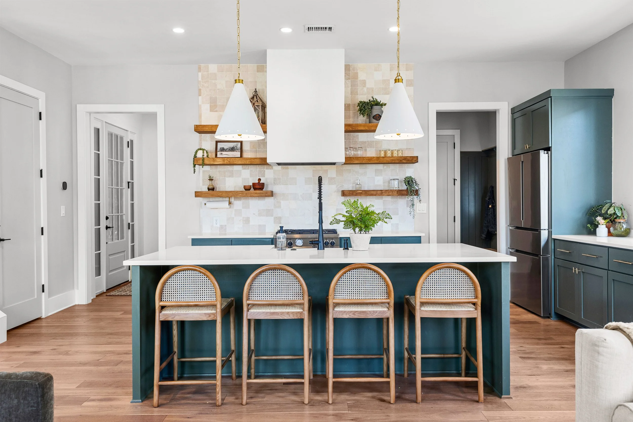 Modern kitchen with white countertops, teal island, wooden bar stools, open shelves with decor, stainless steel refrigerator, and wooden flooring.