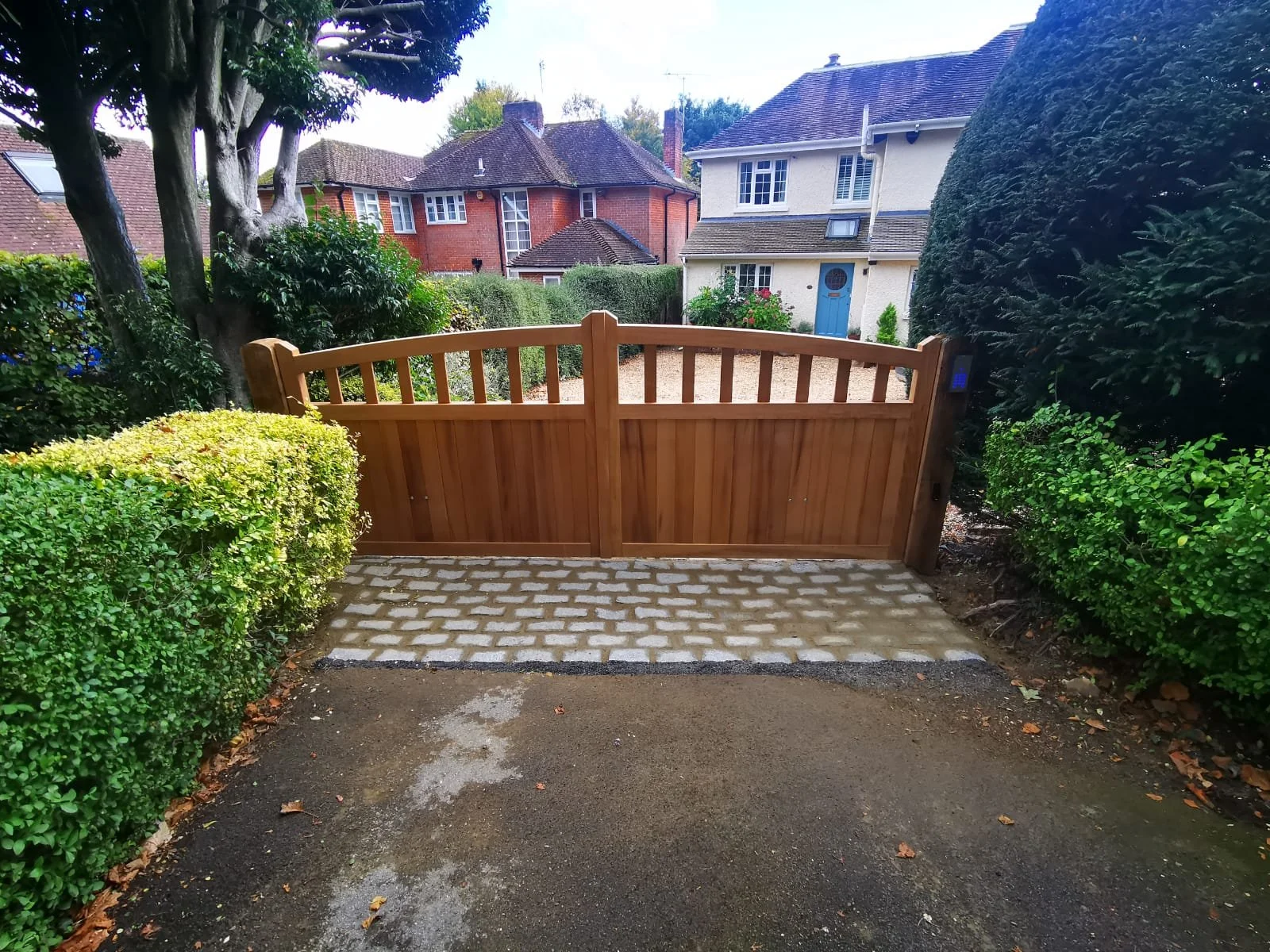 Wooden garden gate with brick pavers leading to a gravel driveway, flanked by green bushes and trees, houses in the background.