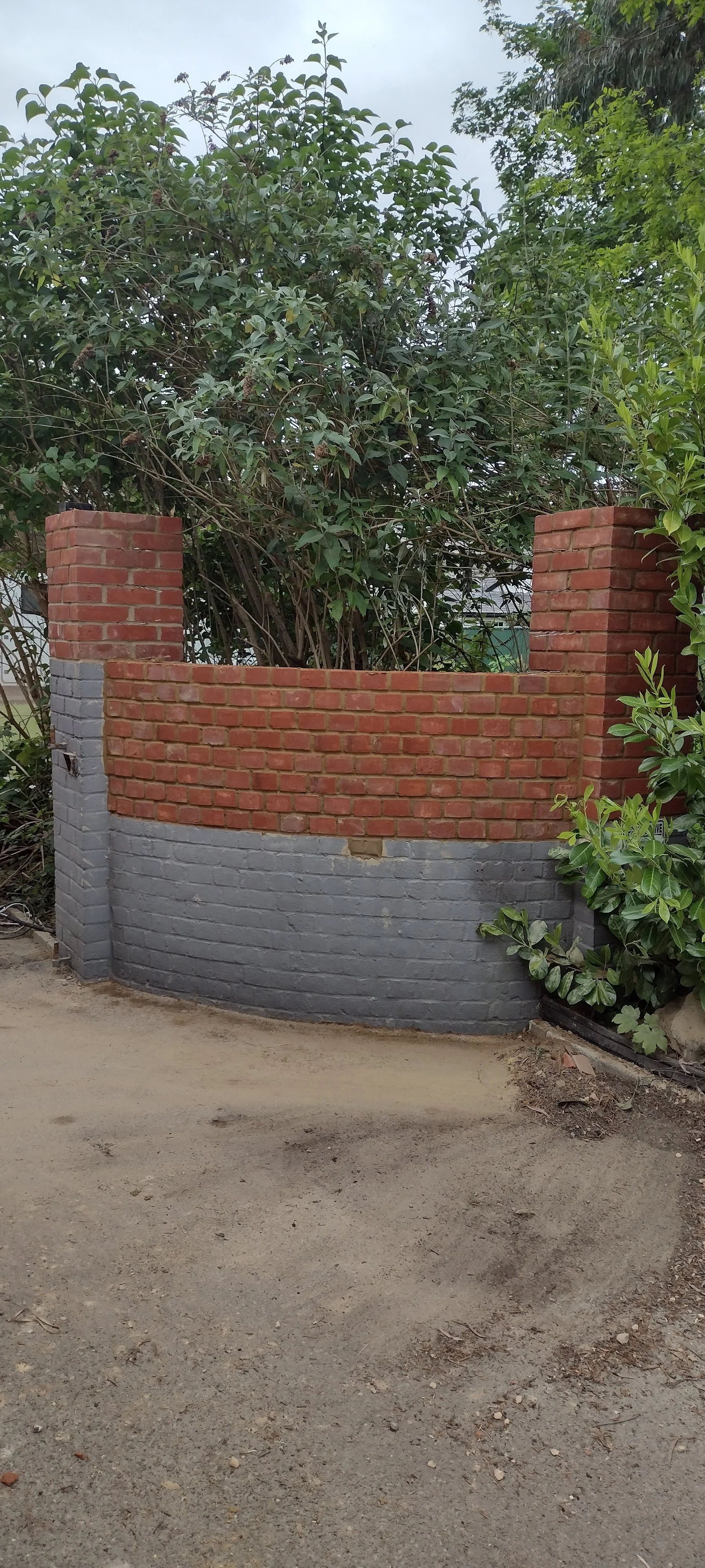 Partial view of a construction site with a curved brick wall, area of dirt ground, and leafy bushes and trees in the background.