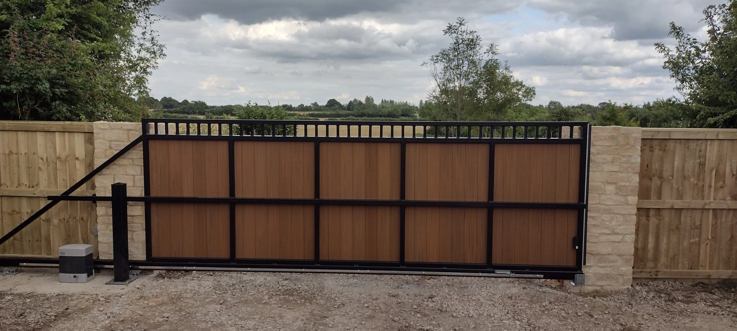 A black and brown wooden gate with a metal frame, set between two stone pillars, in a backyard with a gravel ground and trees in the distance under cloudy sky.